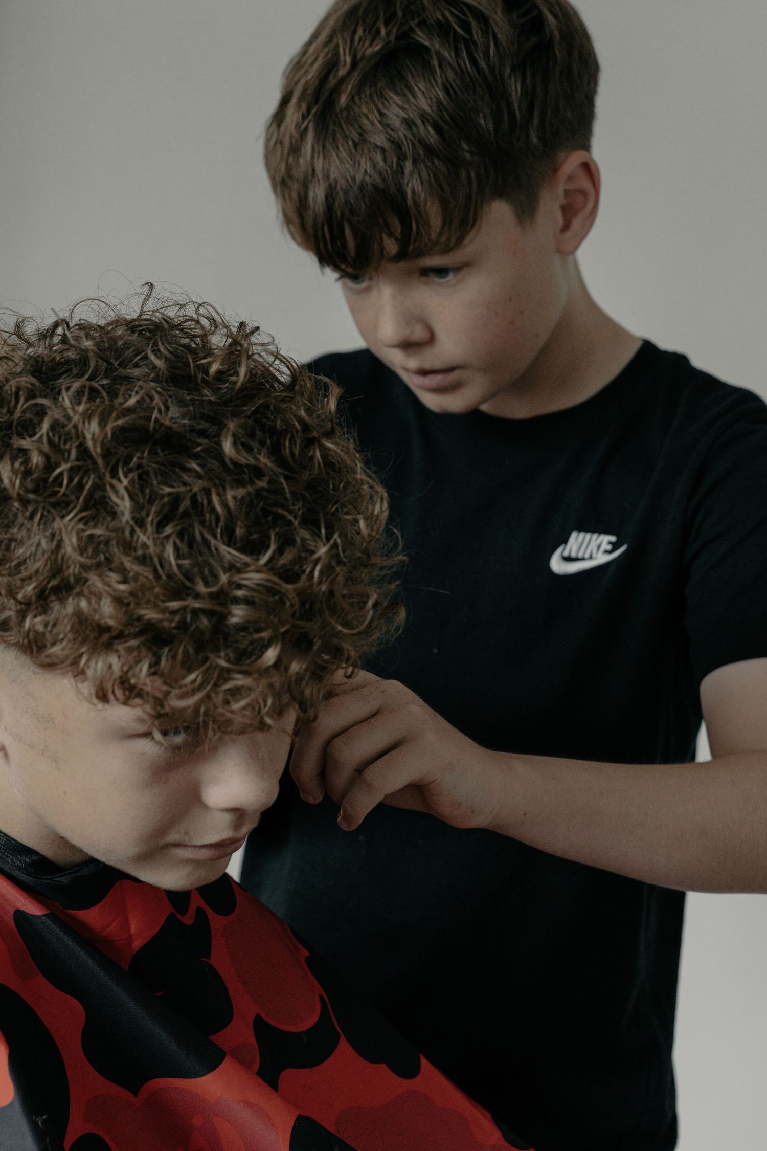 A young boy in a black Nike t-shirt carefully cuts another boys curly hair using hair clippers.