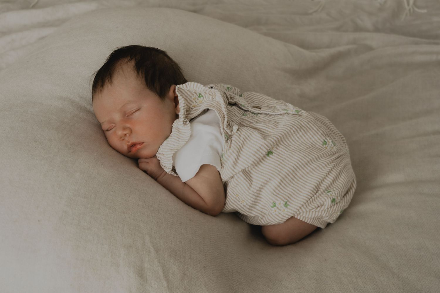 Peaceful newborn baby sleeping curled up on a soft beige linen surface, wearing a striped romper.