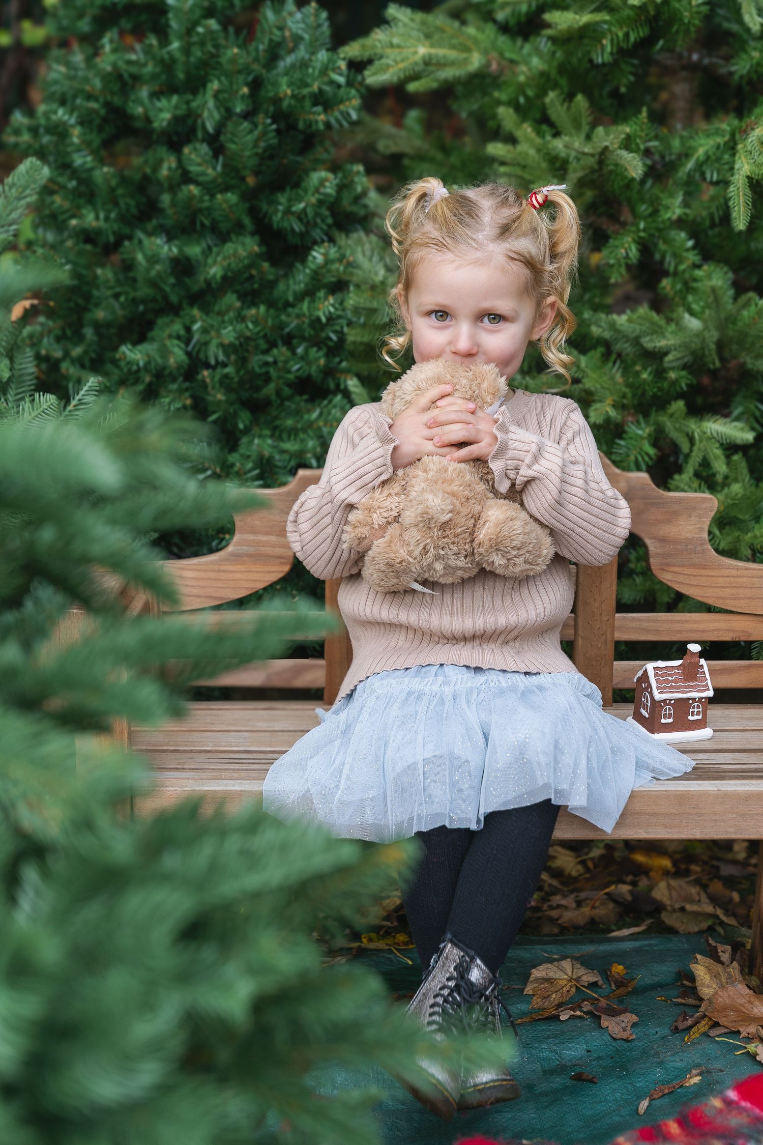 Cute blonde toddler girl hugging teddy bear on wooden bench surrounded by evergreen trees.