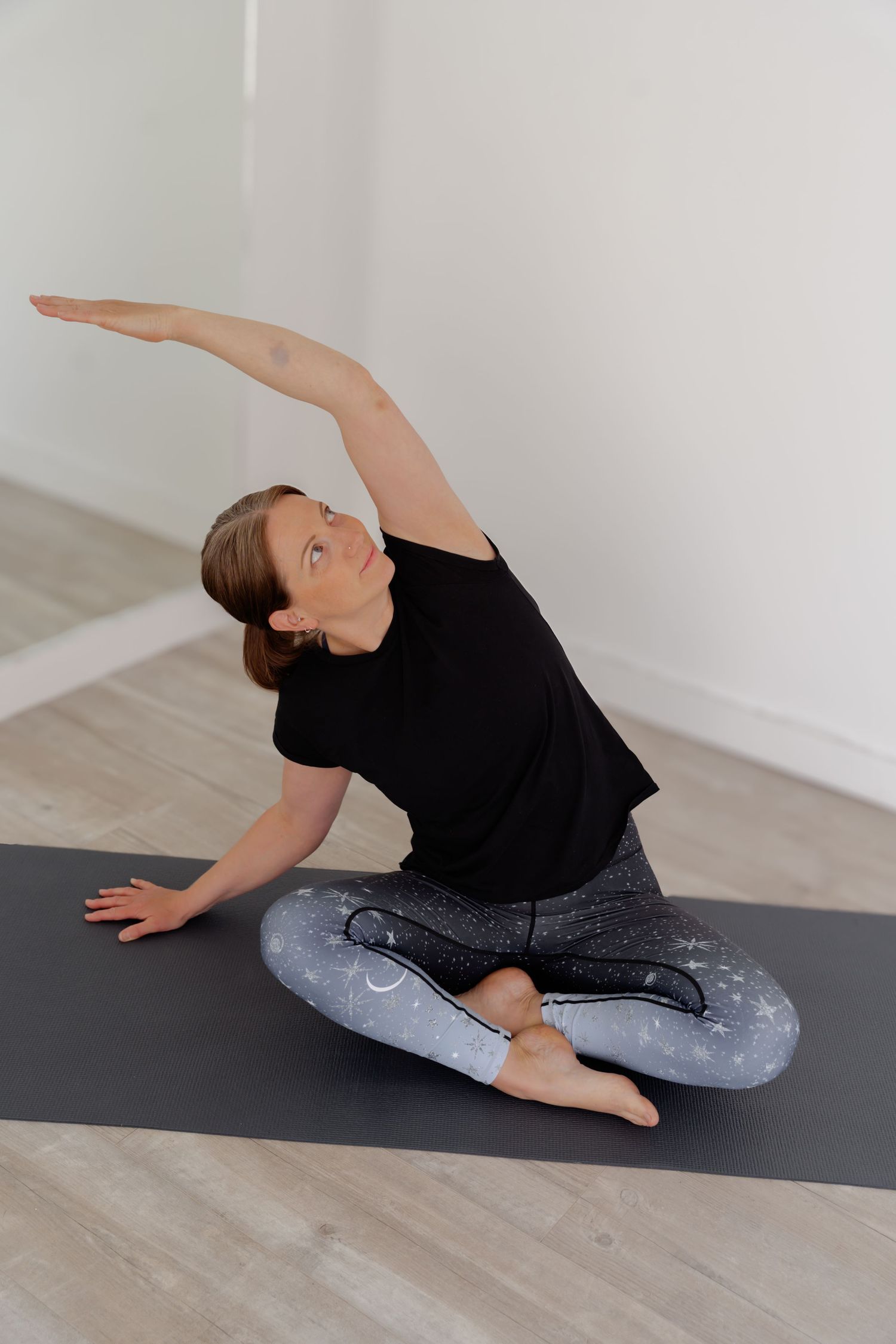 Woman in black top and gray leggings practicing a seated side stretch yoga pose on a dark mat in a bright studio.