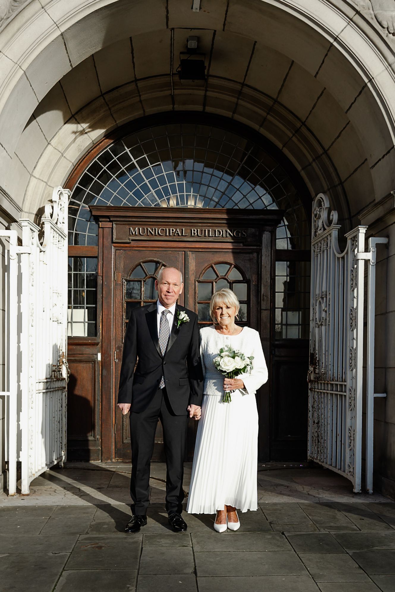 Elegant older couple in wedding attire posing outside a Municipal Building, bride in white dress holding bouquet.