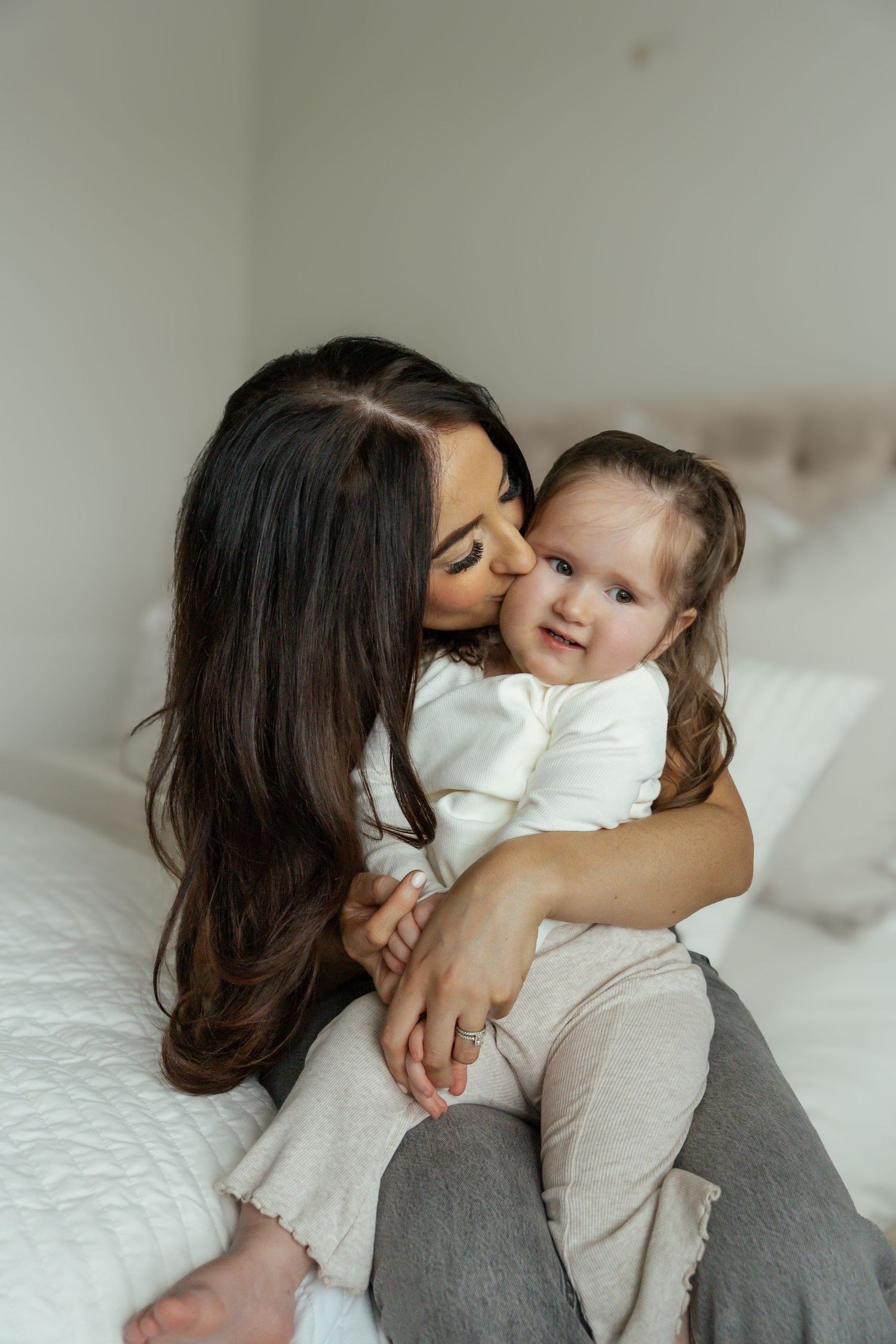 Dark-haired mom tenderly kisses her toddler daughter on the cheek during a cozy bedroom photoshoot.