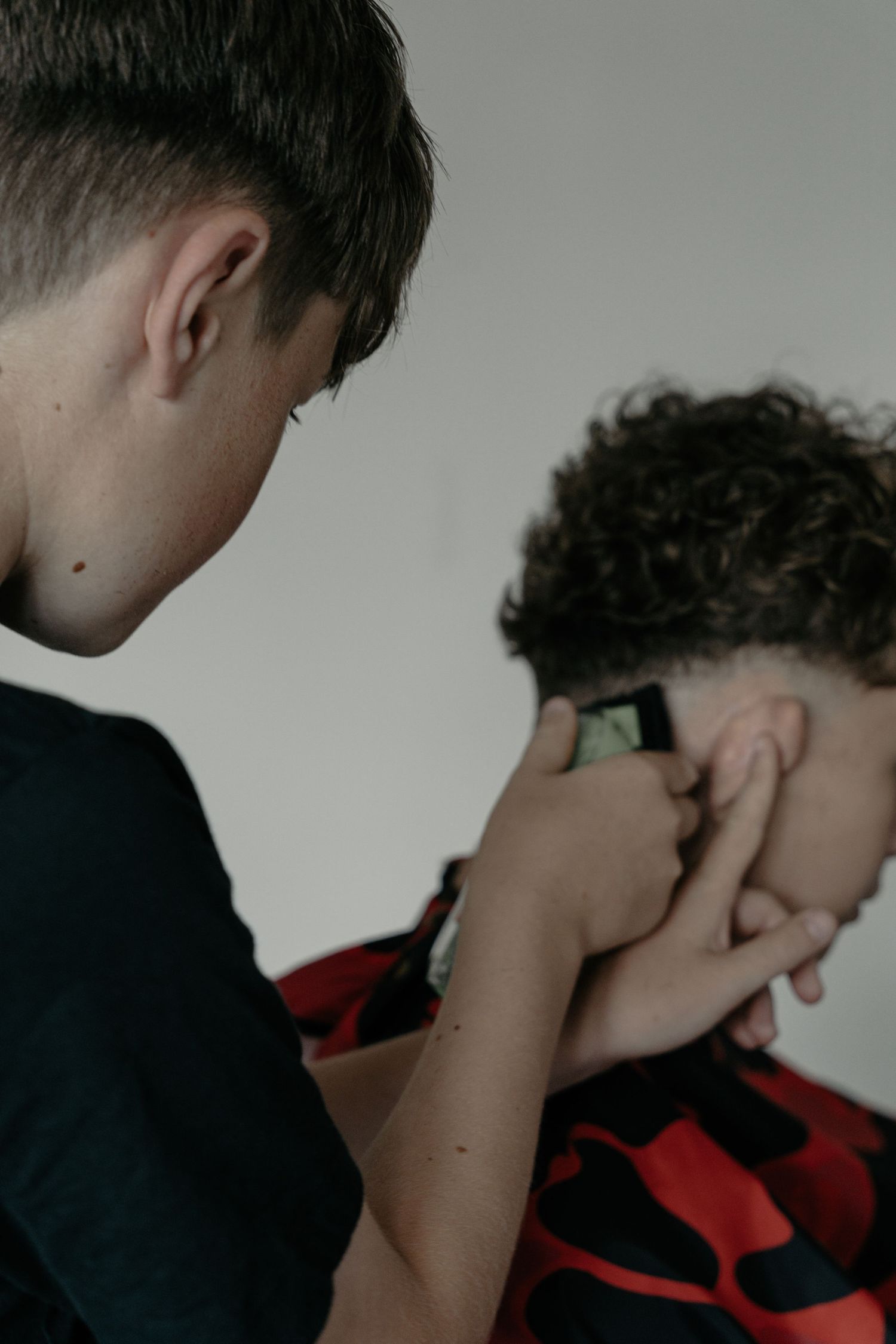 A teenage boy uses electric clippers to trim the neckline of another boys curly hair from behind.