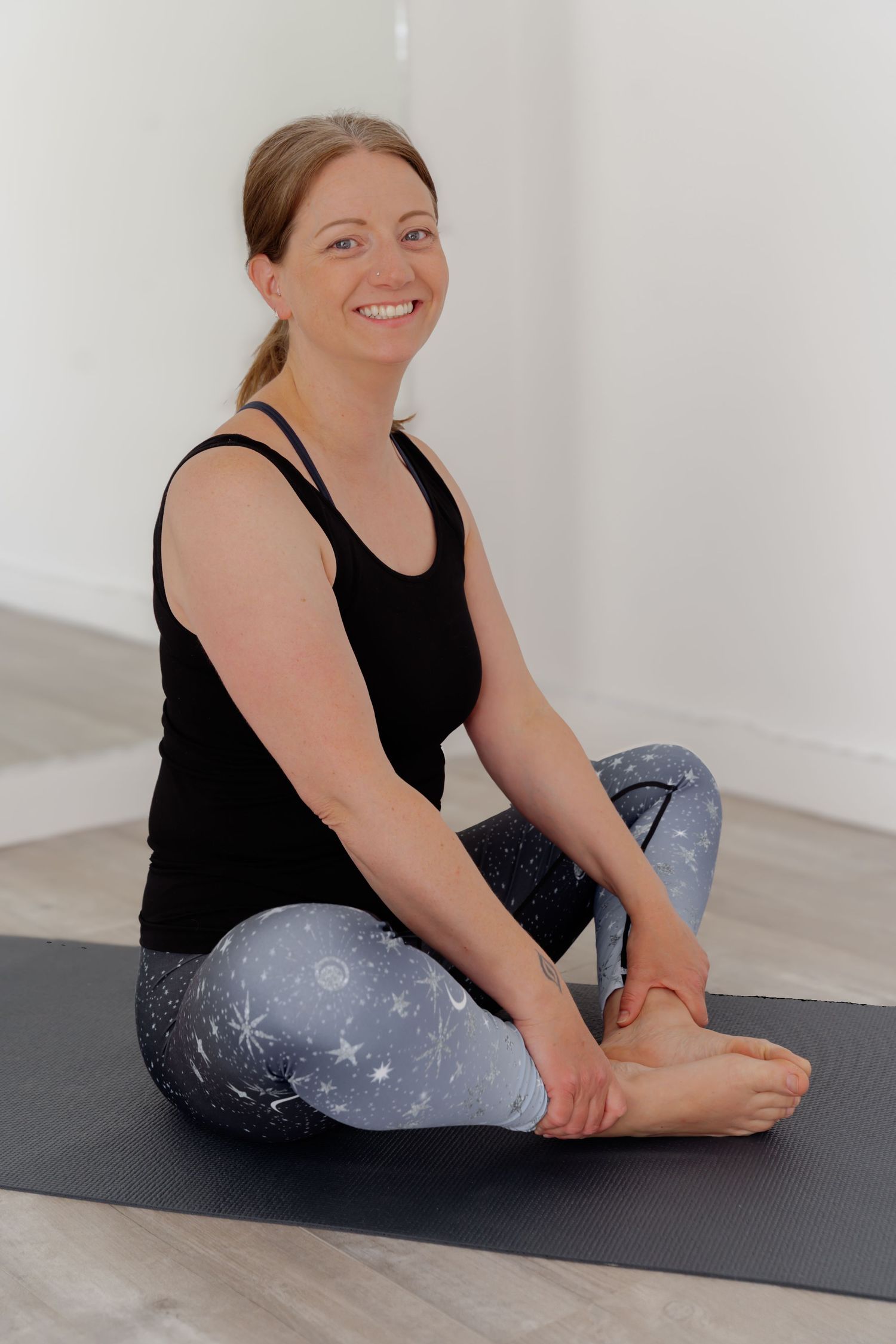 Smiling woman in black tank top and star-print leggings sitting in yoga pose on dark mat in bright studio.