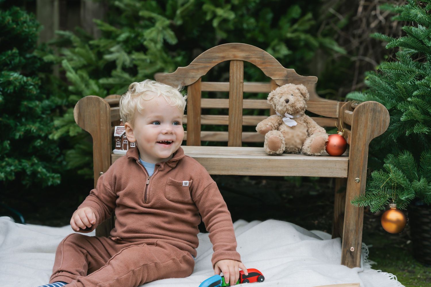 Smiling toddler in brown outfit sits on white blanket beside wooden bench with teddy bear and Christmas ornaments outdoors.