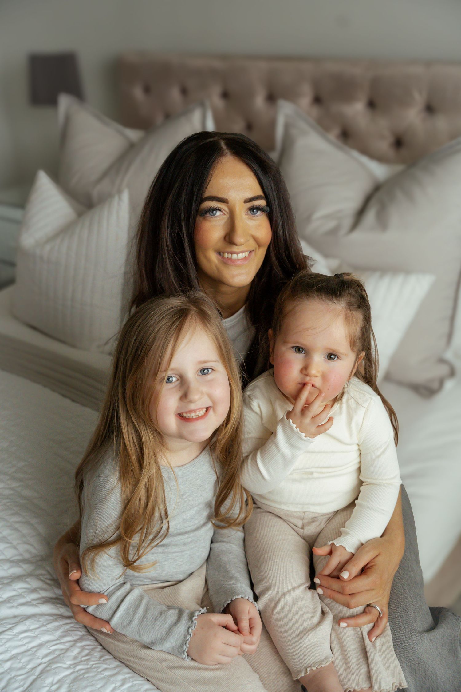 Smiling brunette mother sitting on bed with two young daughters in neutral tones bedroom.