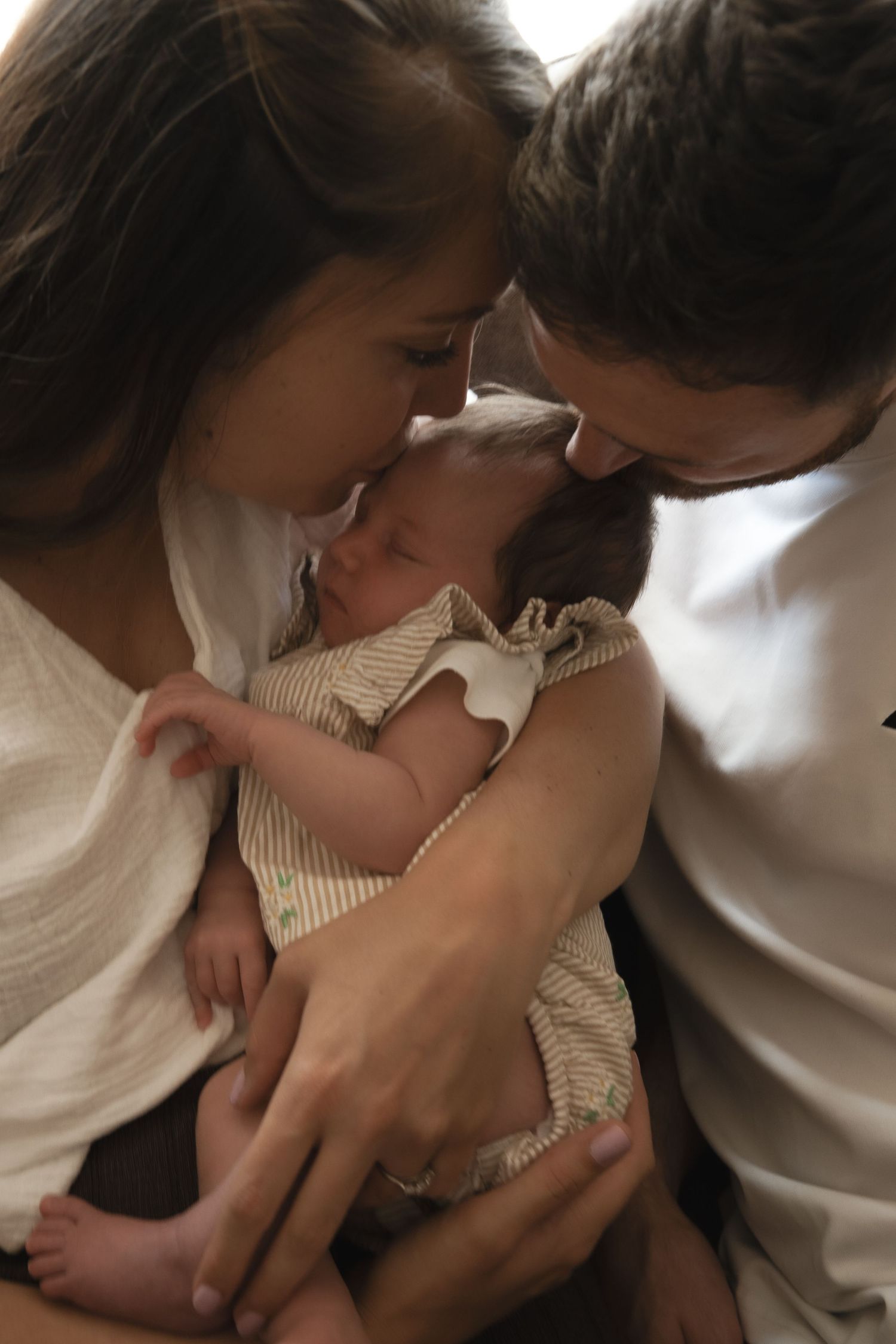 New parents tenderly touch foreheads while cradling their sleeping newborn baby dressed in a striped onesie.