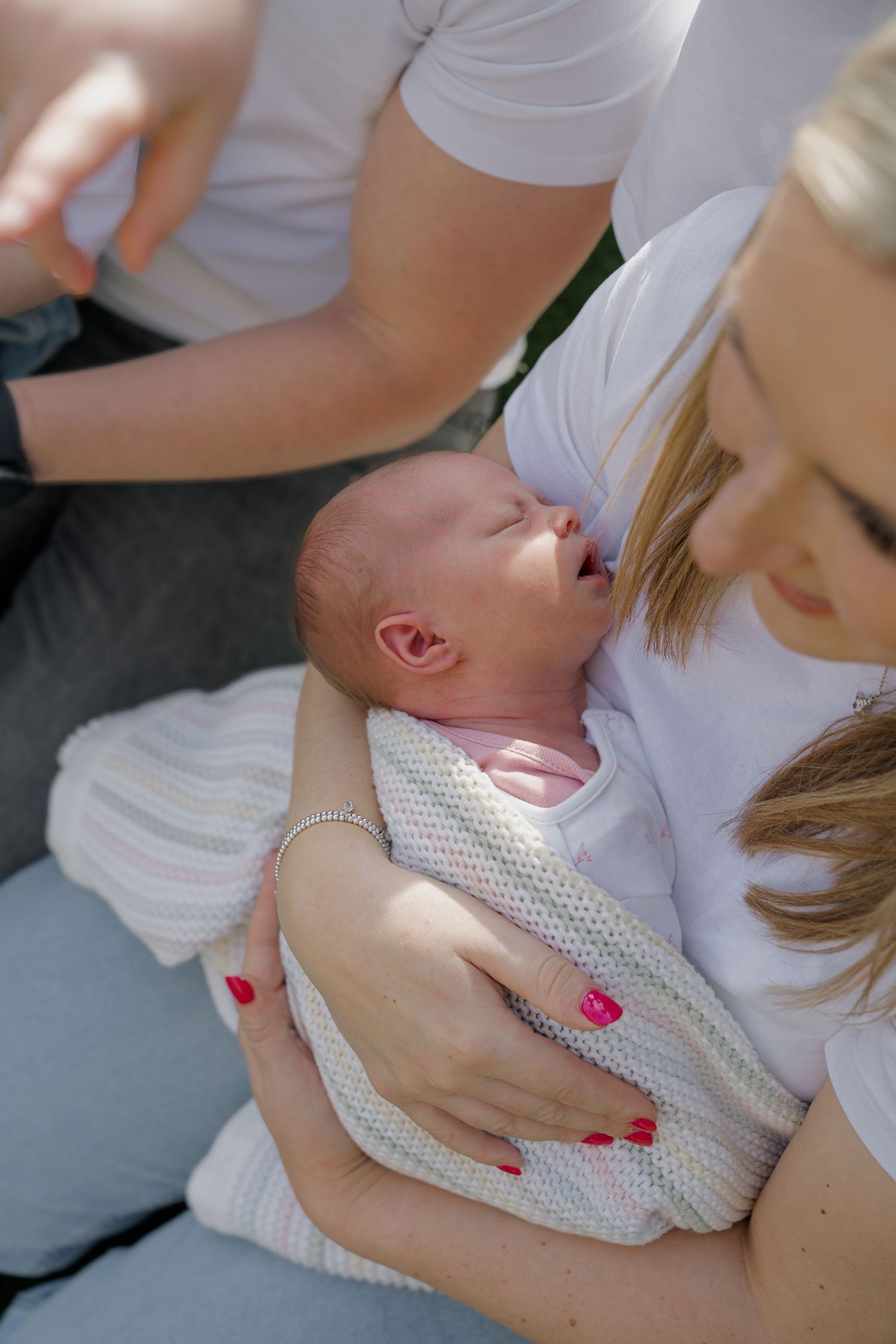 Mother with red nails gently cradling her yawning newborn baby wrapped in a pastel knit blanket outdoors.