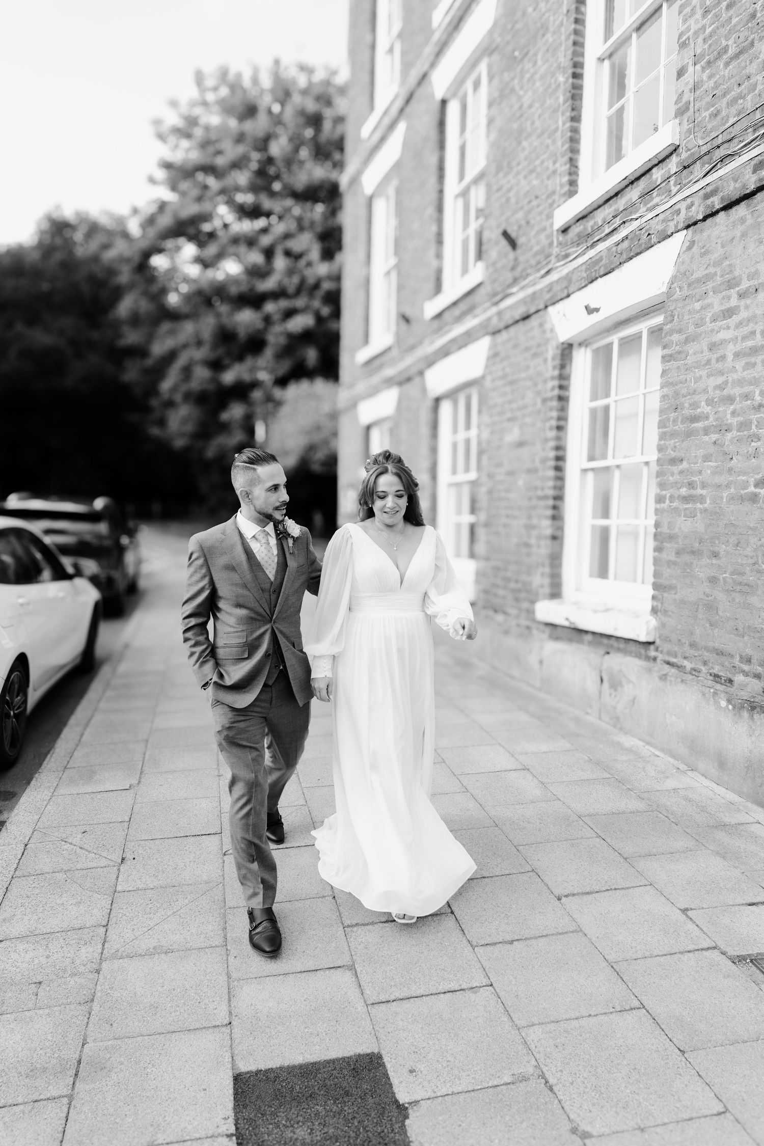 Black and white photo of bride and groom walking hand in hand along a brick building.