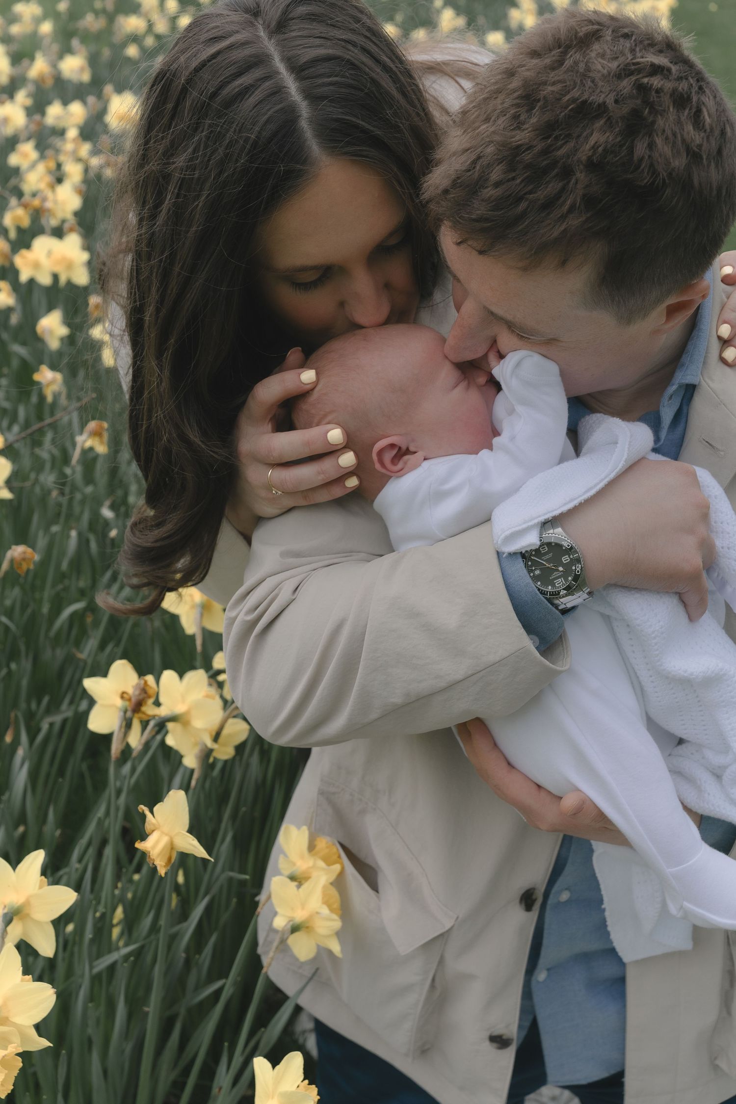 Parents kissing newborn baby together in yellow daffodil flower field in spring.