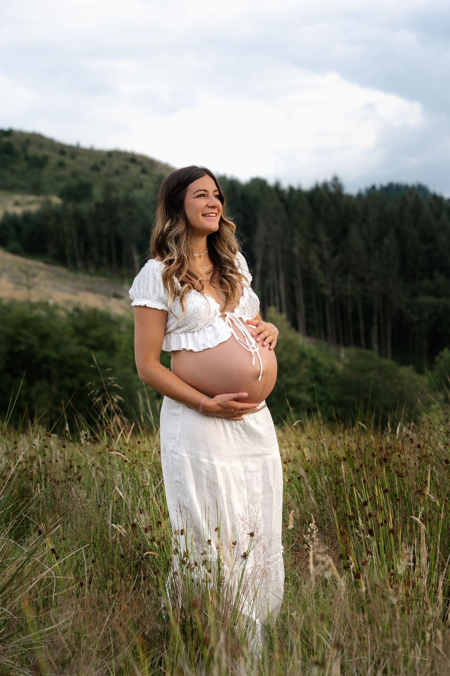 Happy pregnant woman in white dress embracing baby bump outdoors.