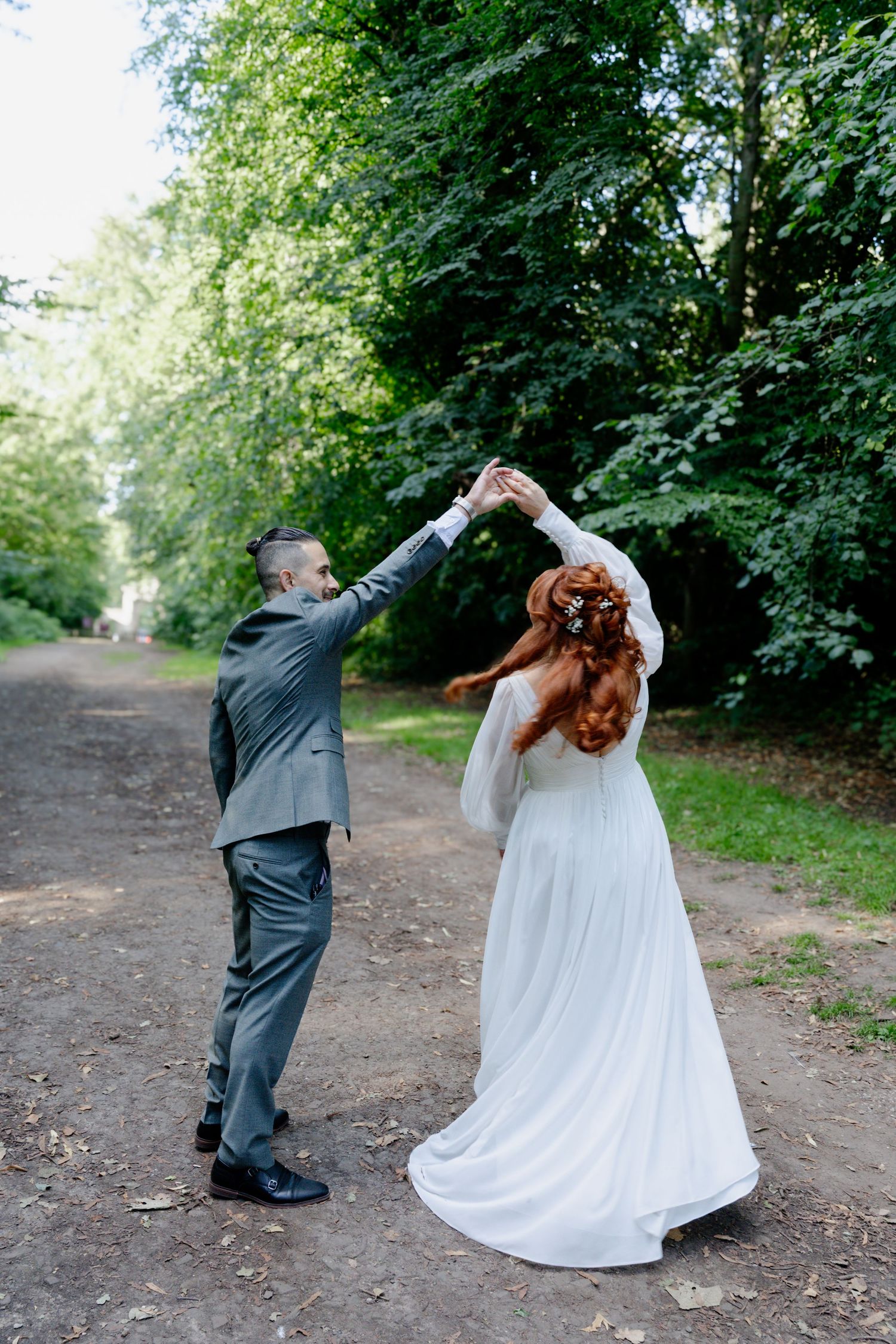 Bride and groom dancing outdoors on a tree-lined path, groom spinning bride in white gown.