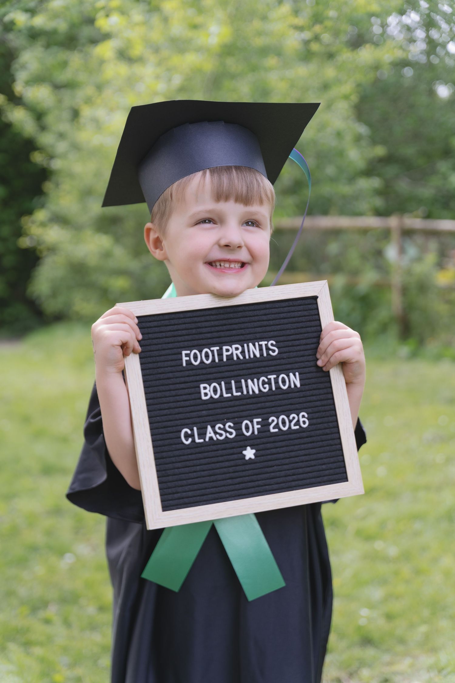 Smiling child in graduation cap and gown holding Footprints Bollington Class of 2026 letterboard sign.
