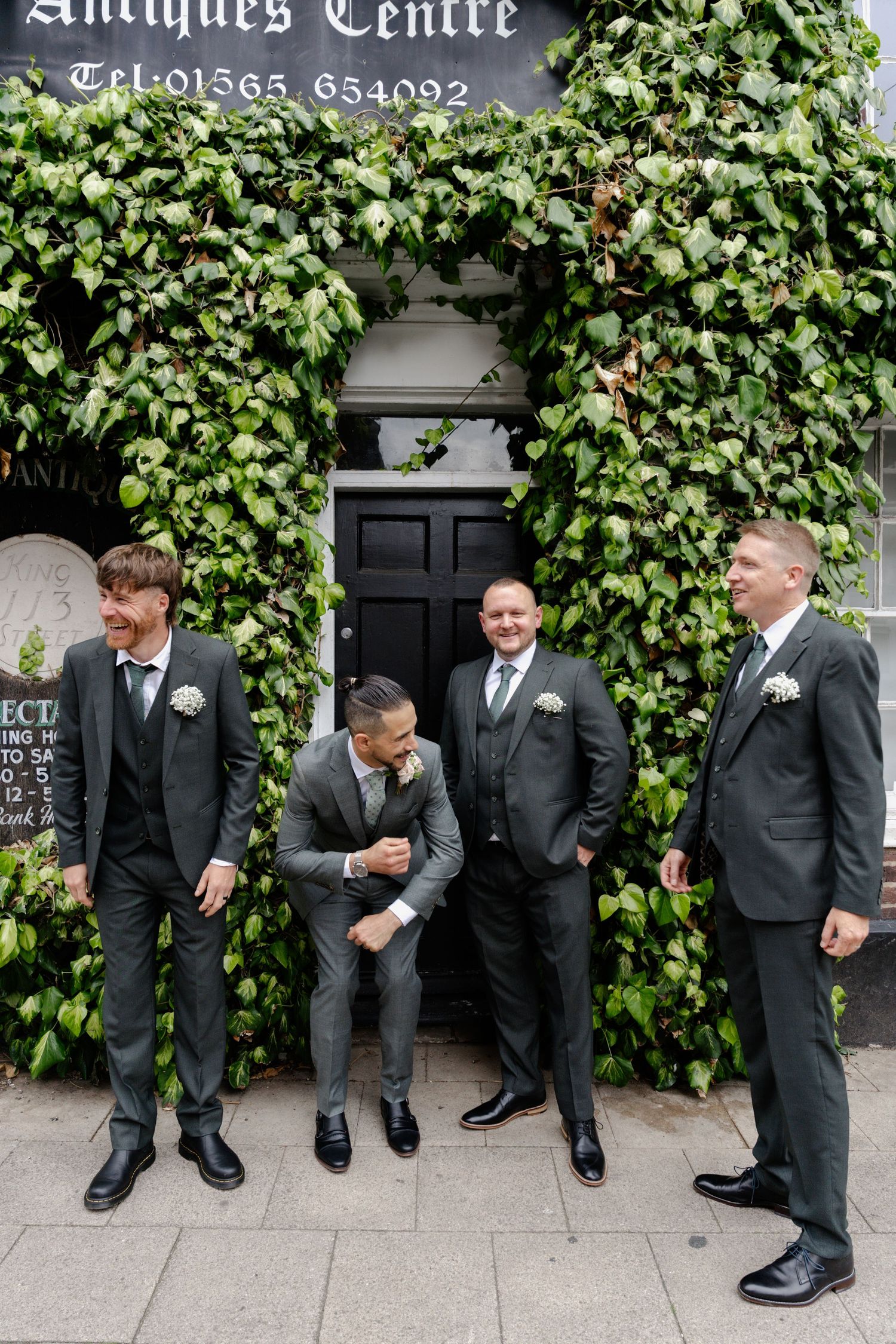 Four groomsmen in grey suits posing outside ivy-covered building with black door at wedding venue.