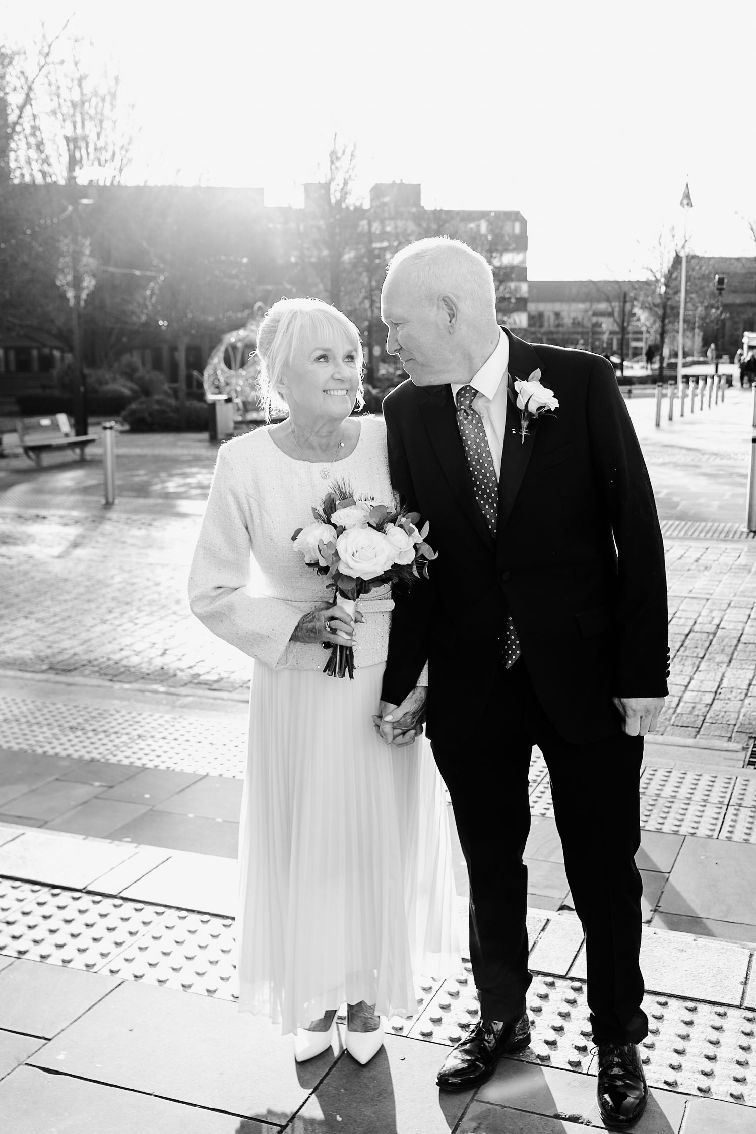 Elderly couple in wedding attire pose outdoors, bride holds bouquet, groom wears suit with boutonniere, black and white photo.