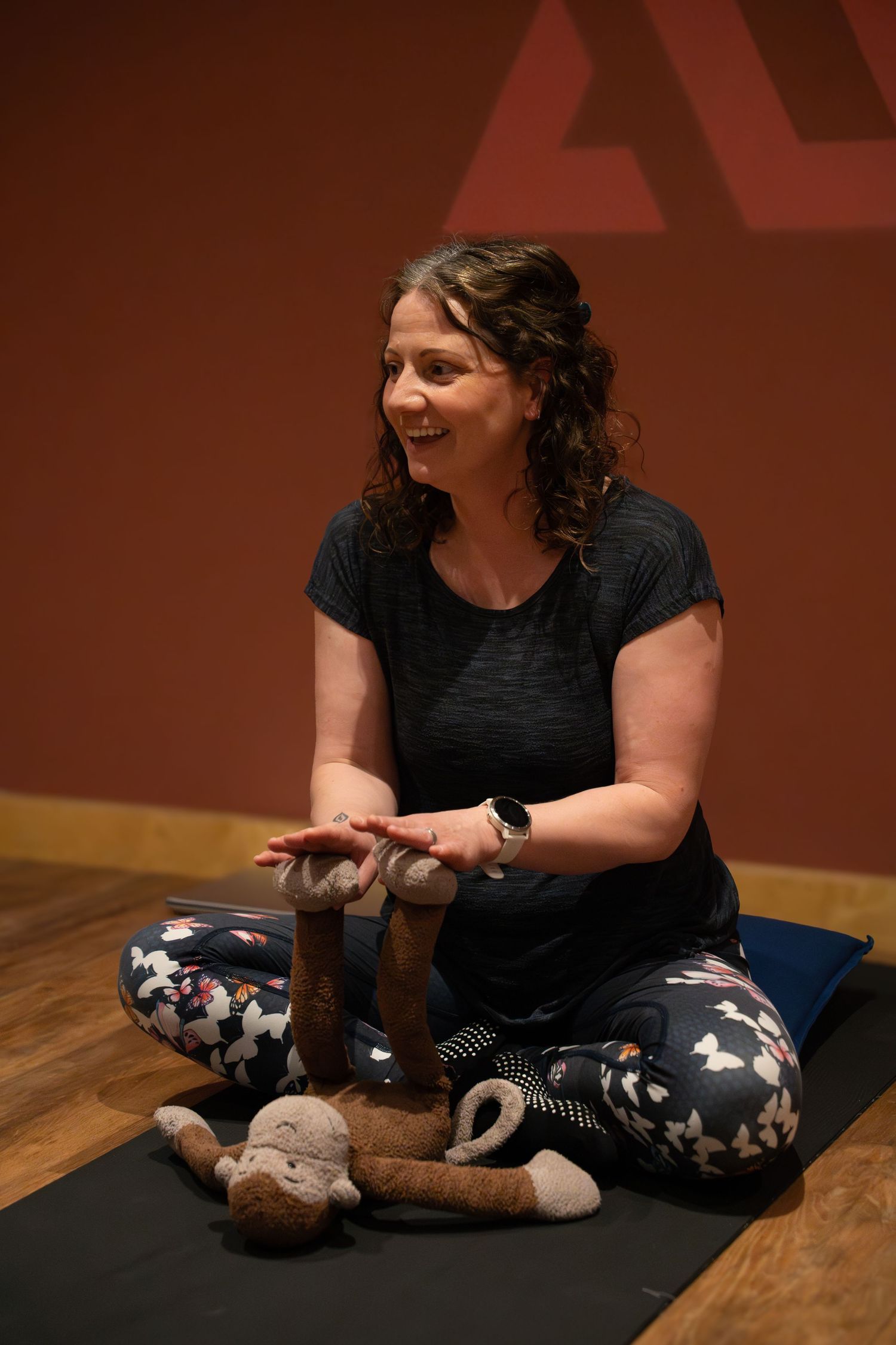 Woman sitting cross-legged on yoga mat with stuffed animals, smiling during a relaxed indoor yoga or mindfulness session.