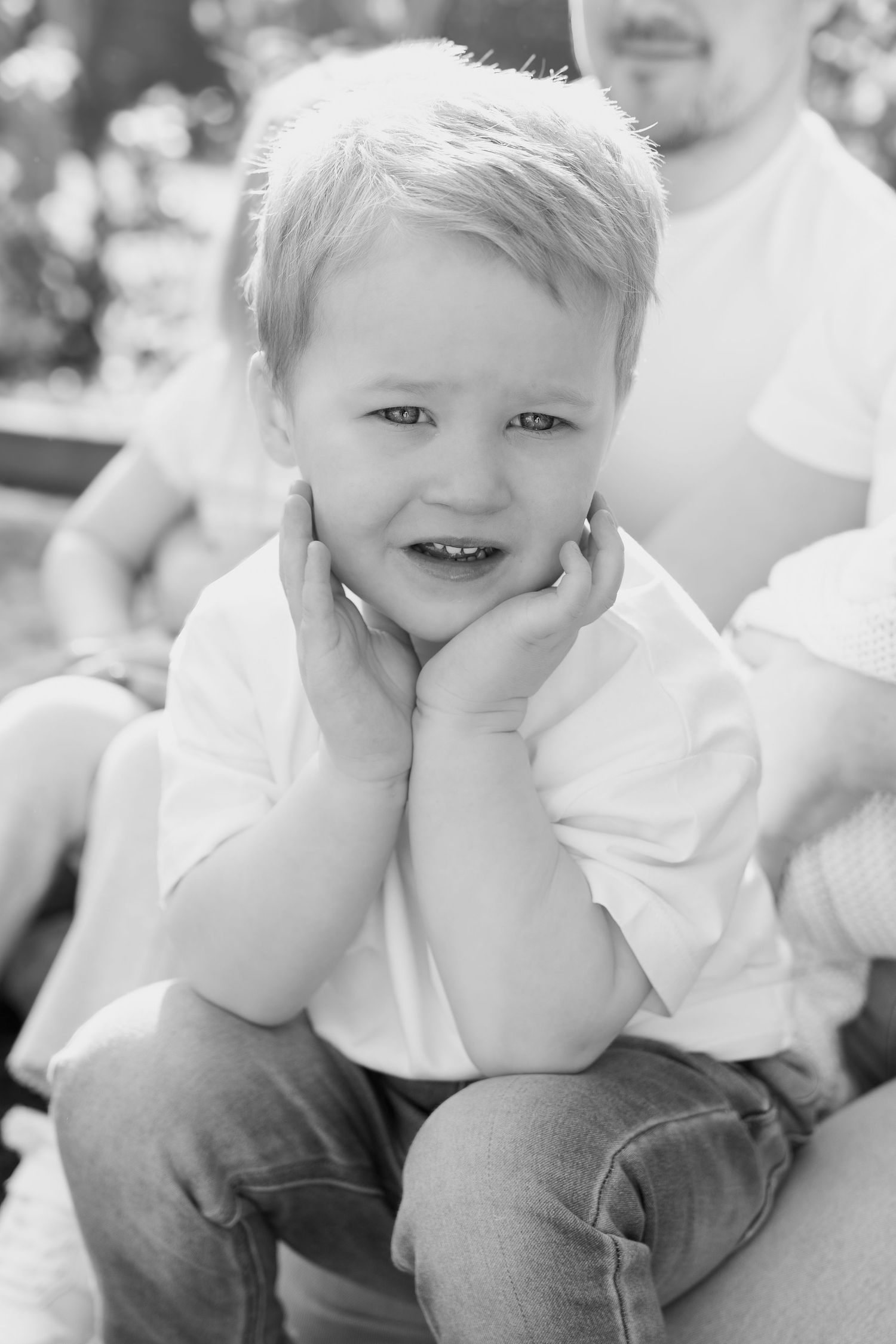 Adorable blonde toddler boy smiling with hands on cheeks in a charming black and white outdoor portrait.