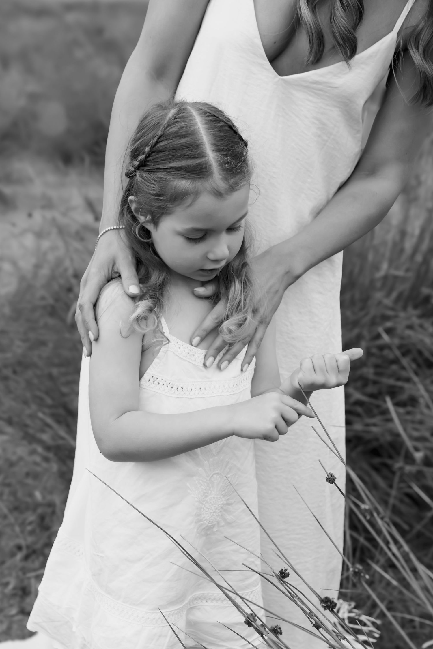 Black and white photo of young girl in white dress examining wildflowers in a field.