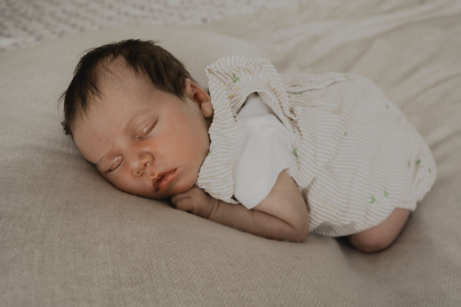 Peaceful newborn baby sleeping on a soft beige blanket wearing a white knit romper.