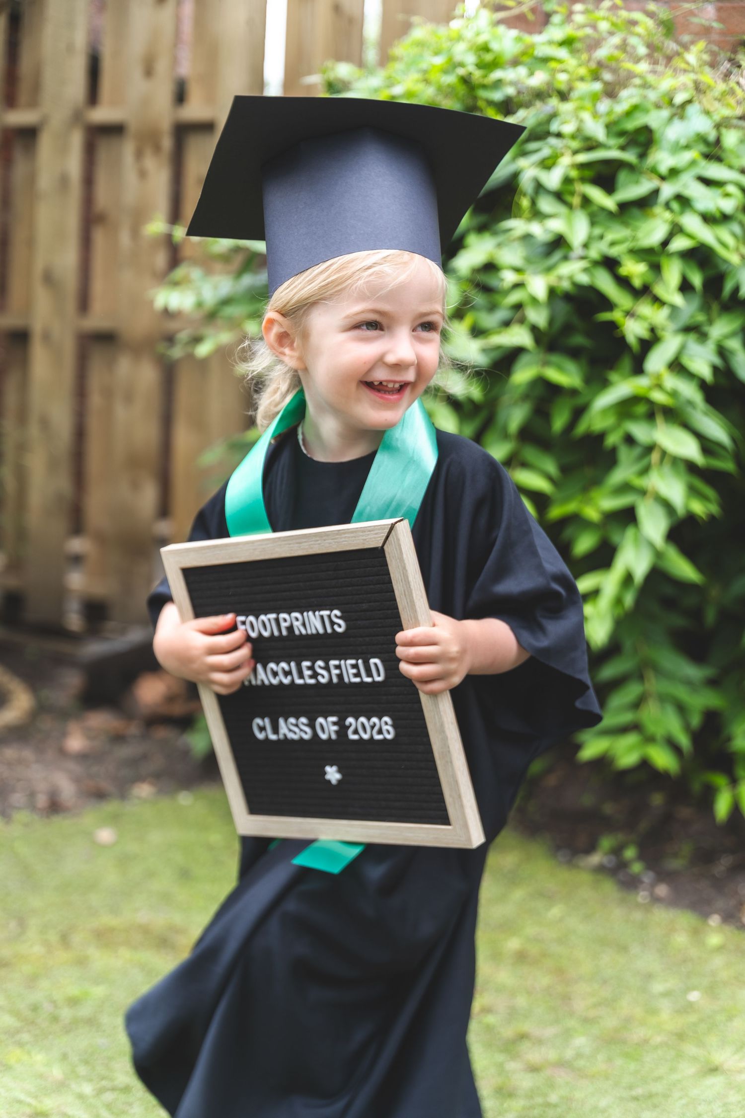 Smiling toddler in black graduation gown and cap holds a letter board reading Footprints Macclesfield Class of 2026.
