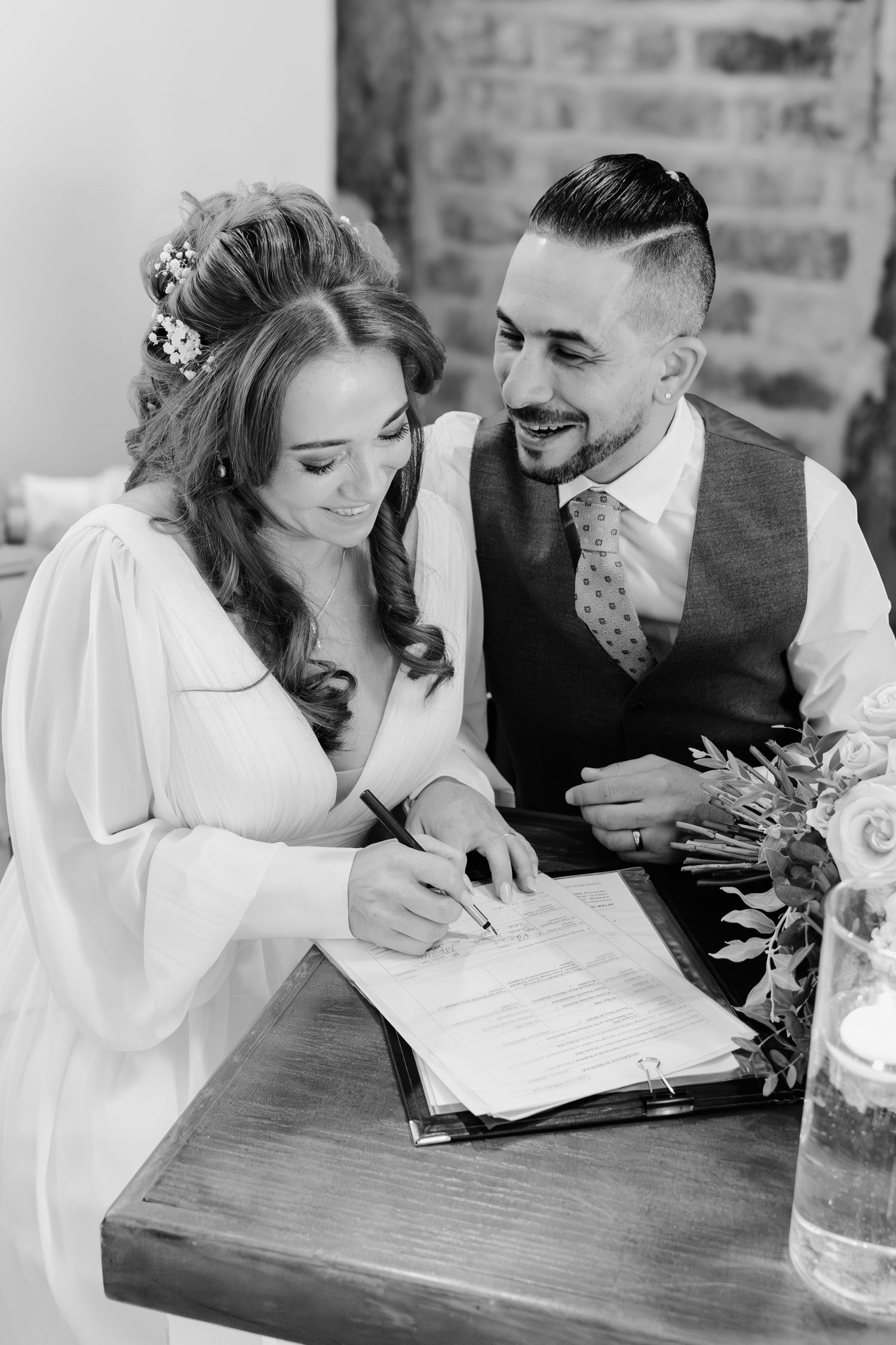Bride and groom smiling while signing wedding register at wooden table in black and white photo.