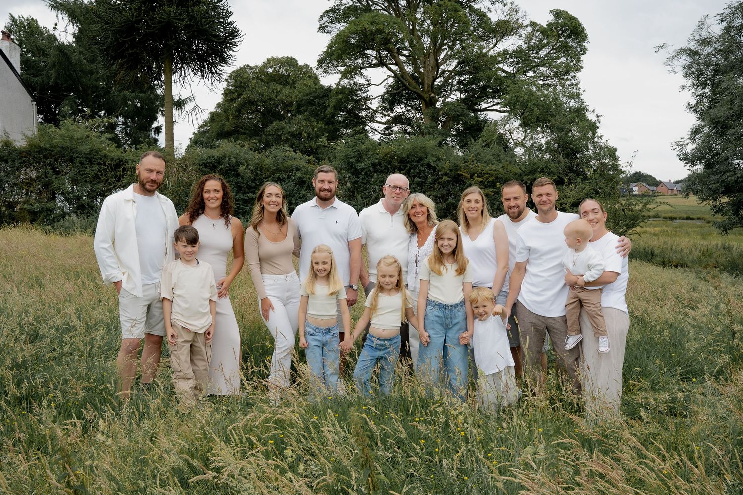 Large extended family group portrait in white outfits standing in lush green meadow outdoors.