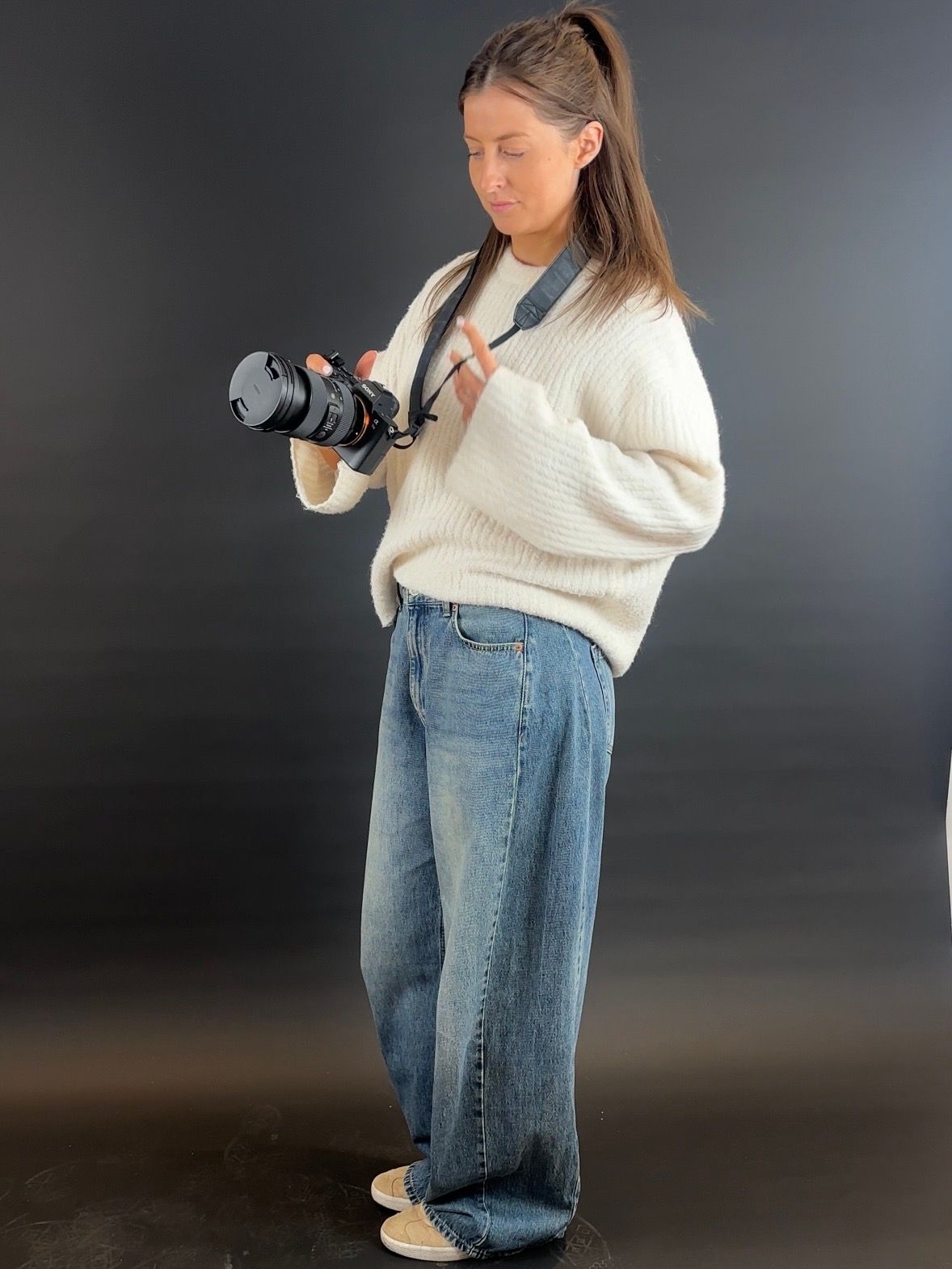 Female photographer in cream sweater and jeans reviewing images on camera against dark background.
