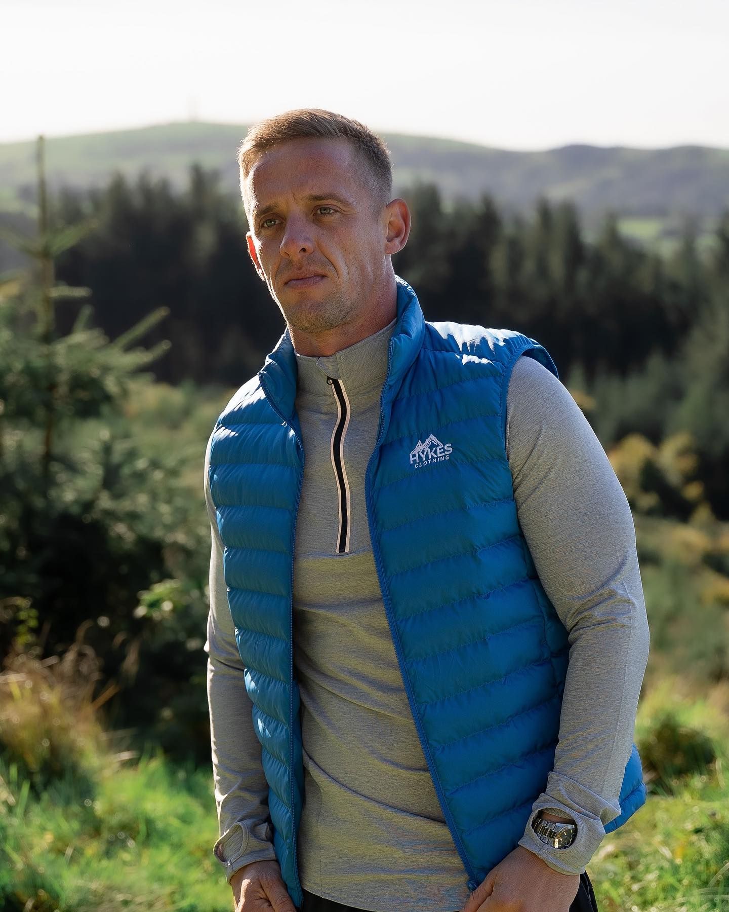 Man wearing a blue puffer vest and grey base layer standing outdoors with green hills and forest in background.