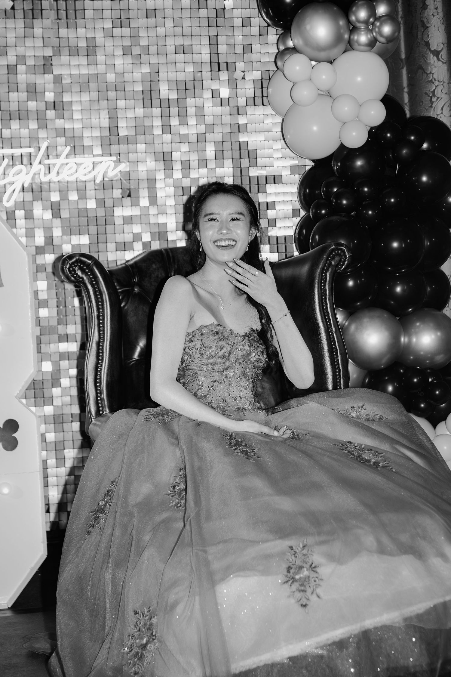 Young woman in elegant ball gown smiles while seated in armchair at 18th birthday celebration with balloon decorations.