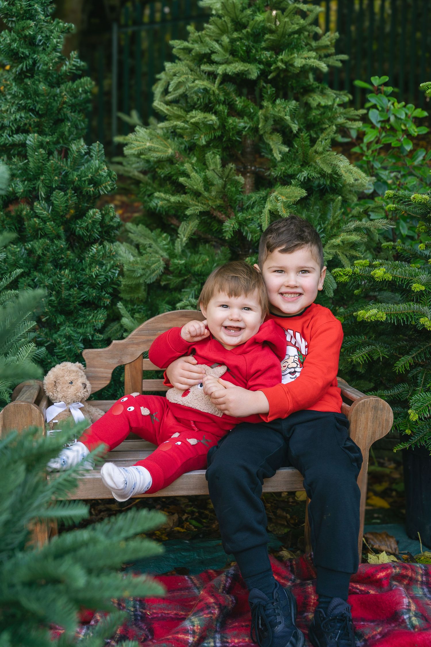 Two happy children in red outfits sit on a wooden bench surrounded by lush green Christmas trees outdoors.