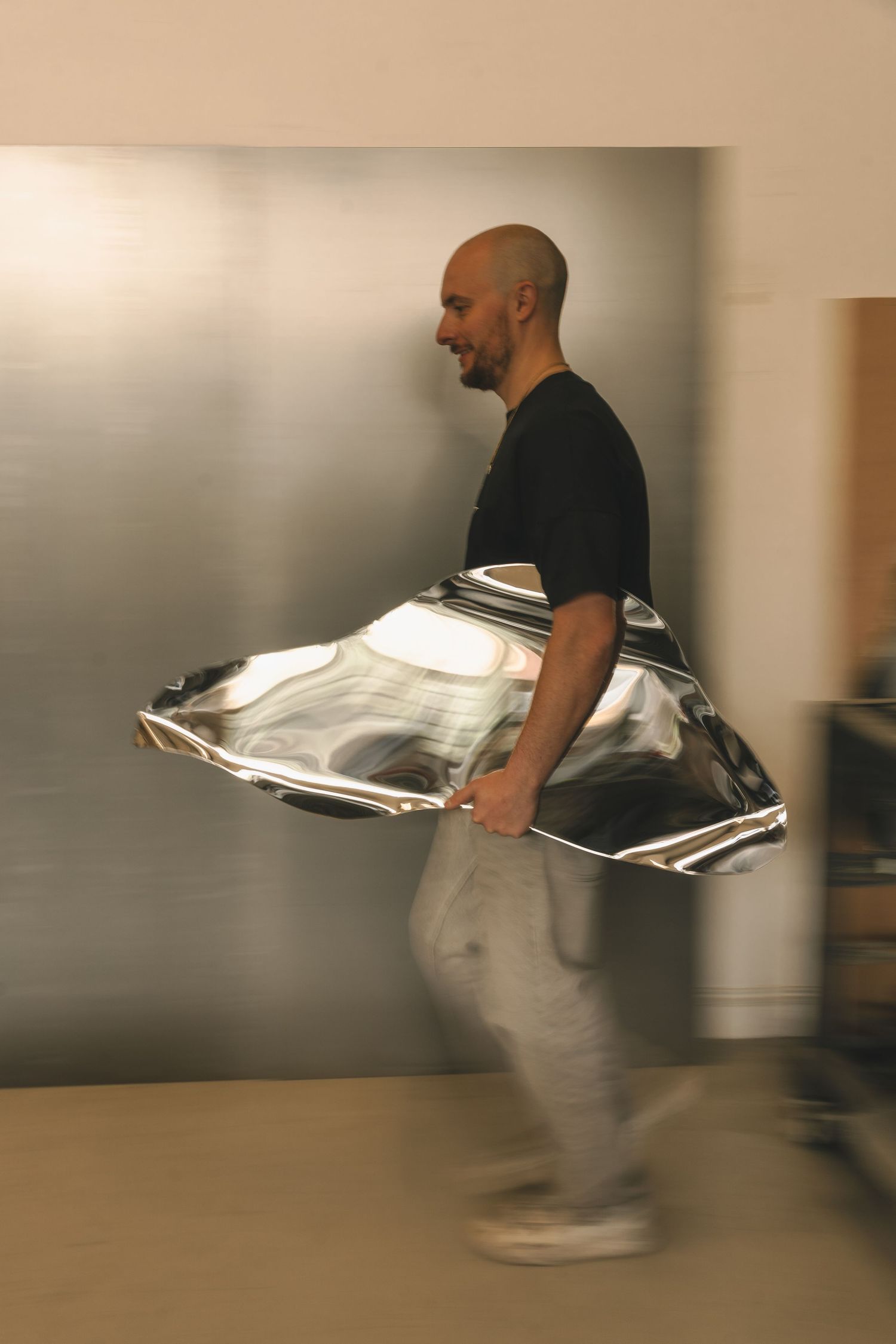 A bald man in a black t-shirt carries a large polished silver metallic object through a workshop.