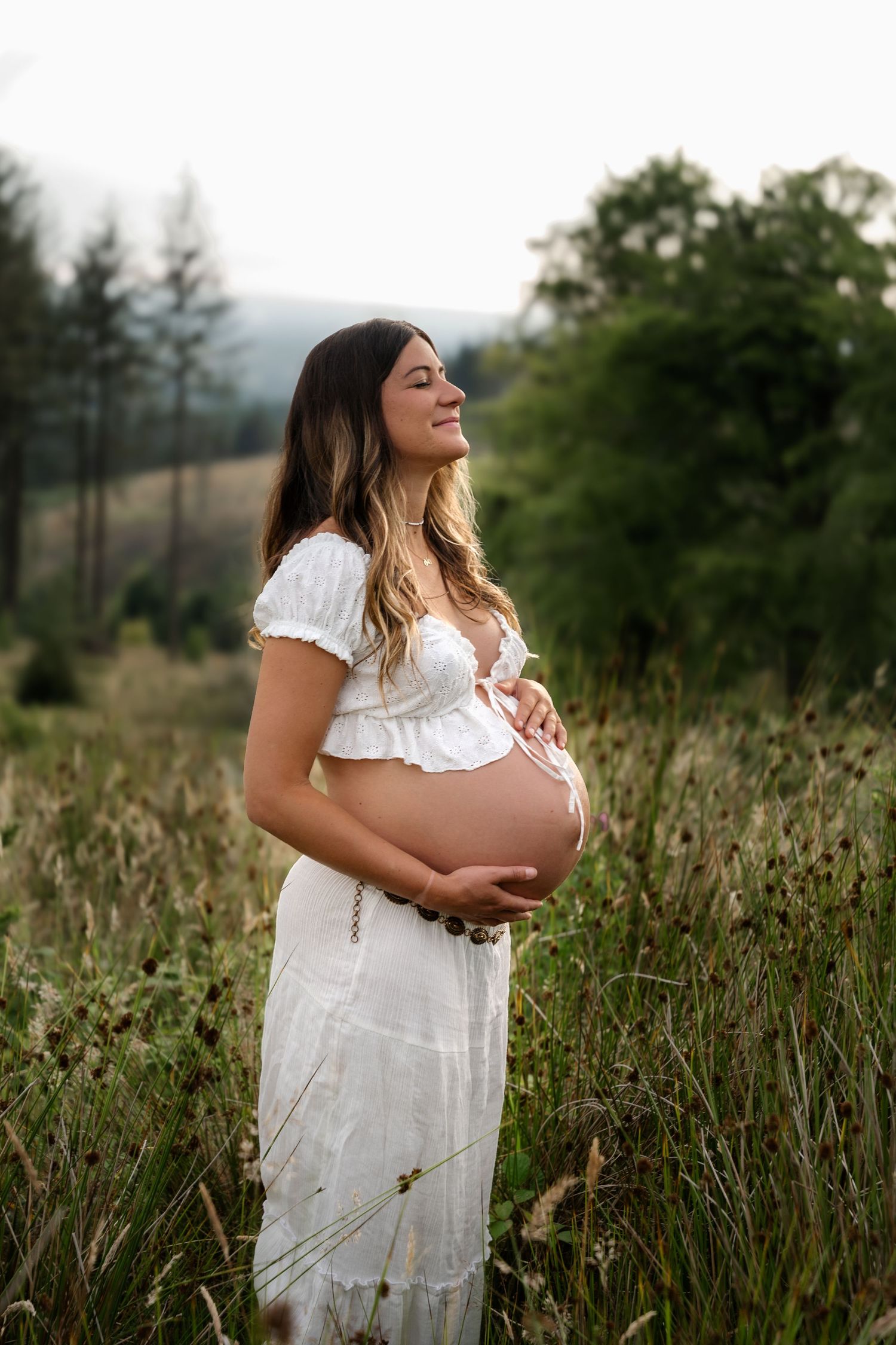 Pregnant woman in white dress holding baby bump in a lush green field.