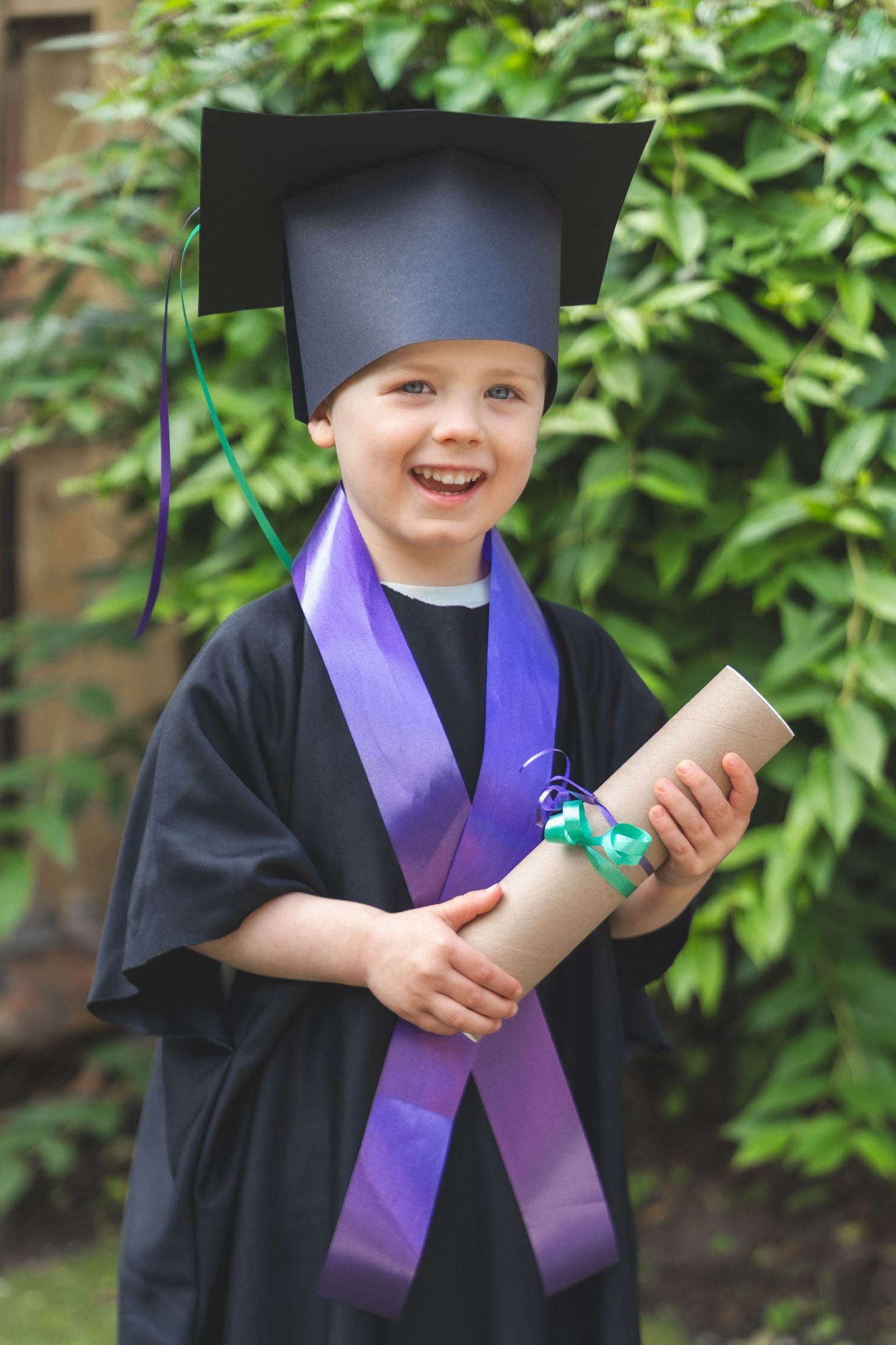 A smiling young child wearing a black graduation gown, mortarboard cap, and purple sash, holding a diploma outdoors.