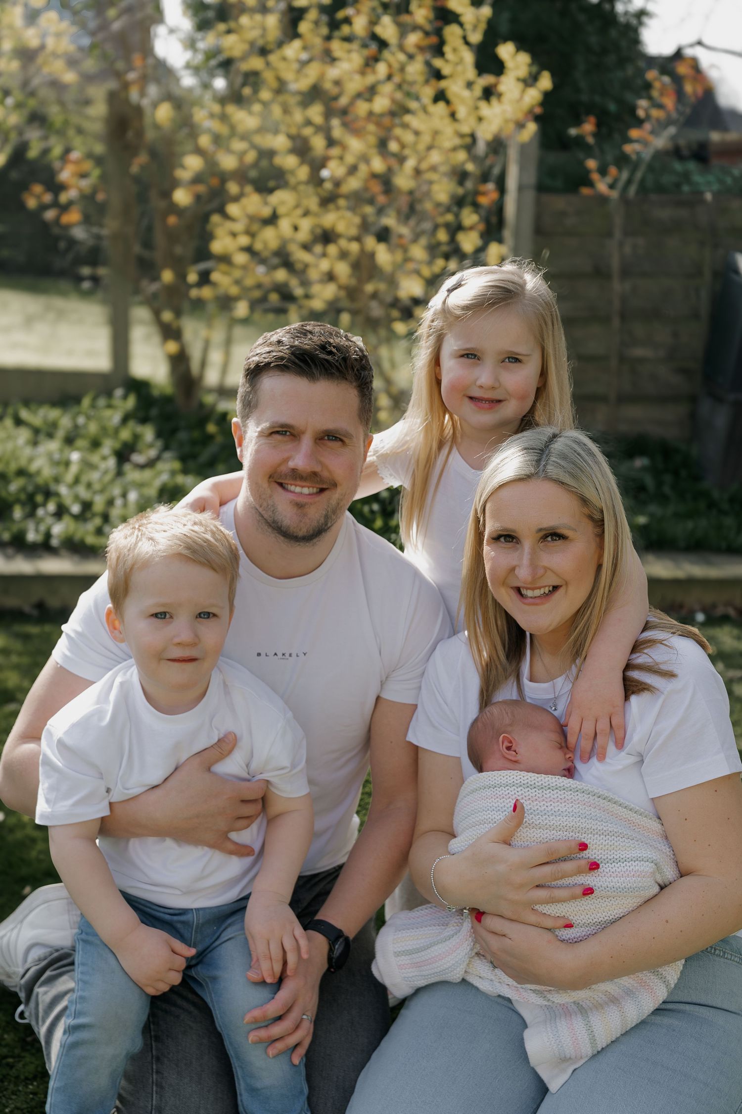 Smiling family portrait with father, mother holding newborn, young son and daughter in a bright garden setting.