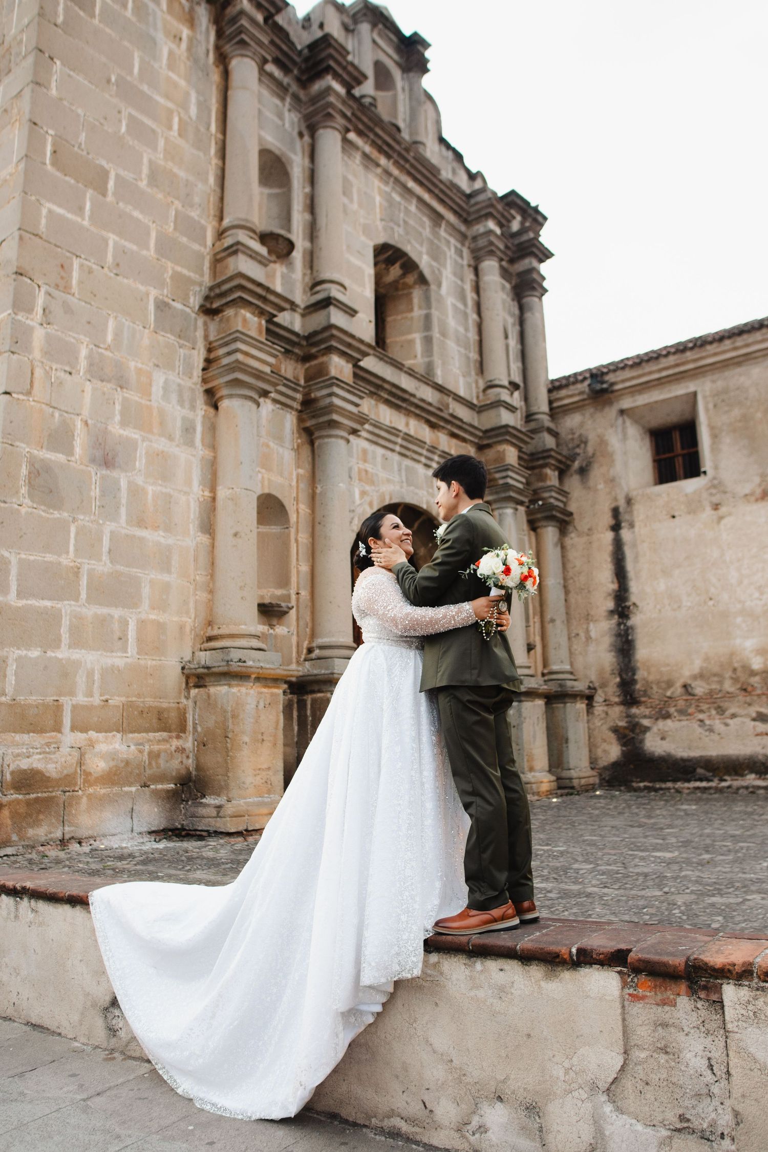 A couple in wedding attire embraces in front of an ornate stone church facade with colonial architecture.