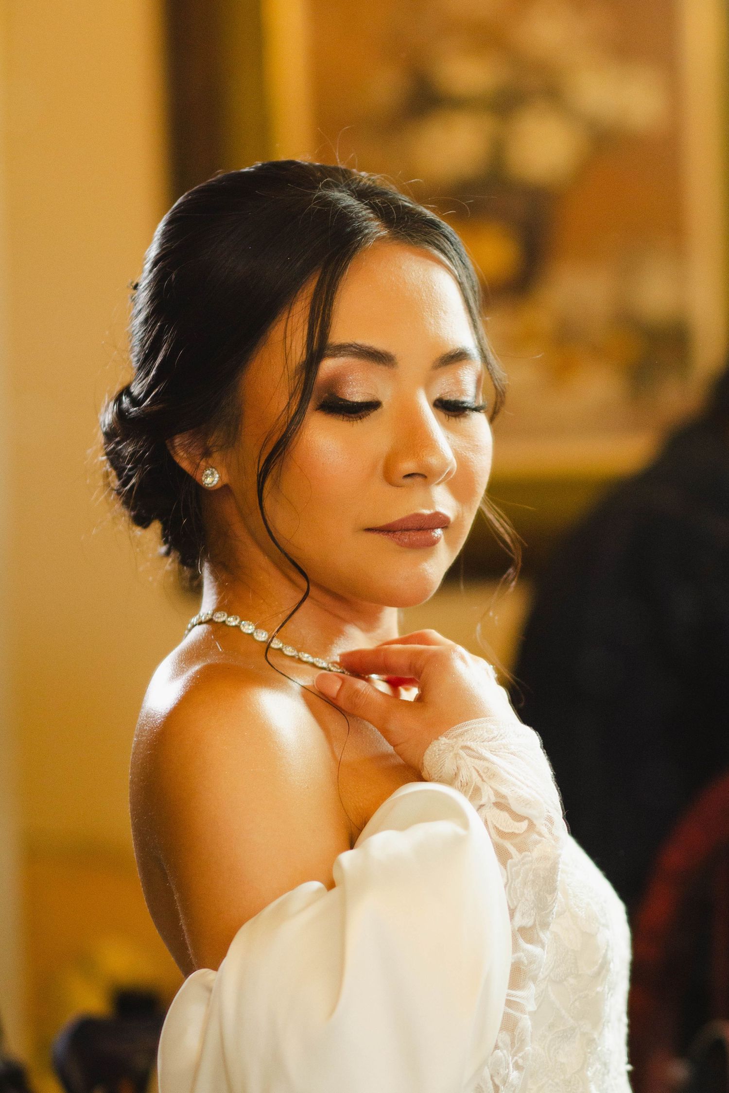 A bride in an off-shoulder wedding dress with pearl necklace poses with eyes closed in warm lighting.