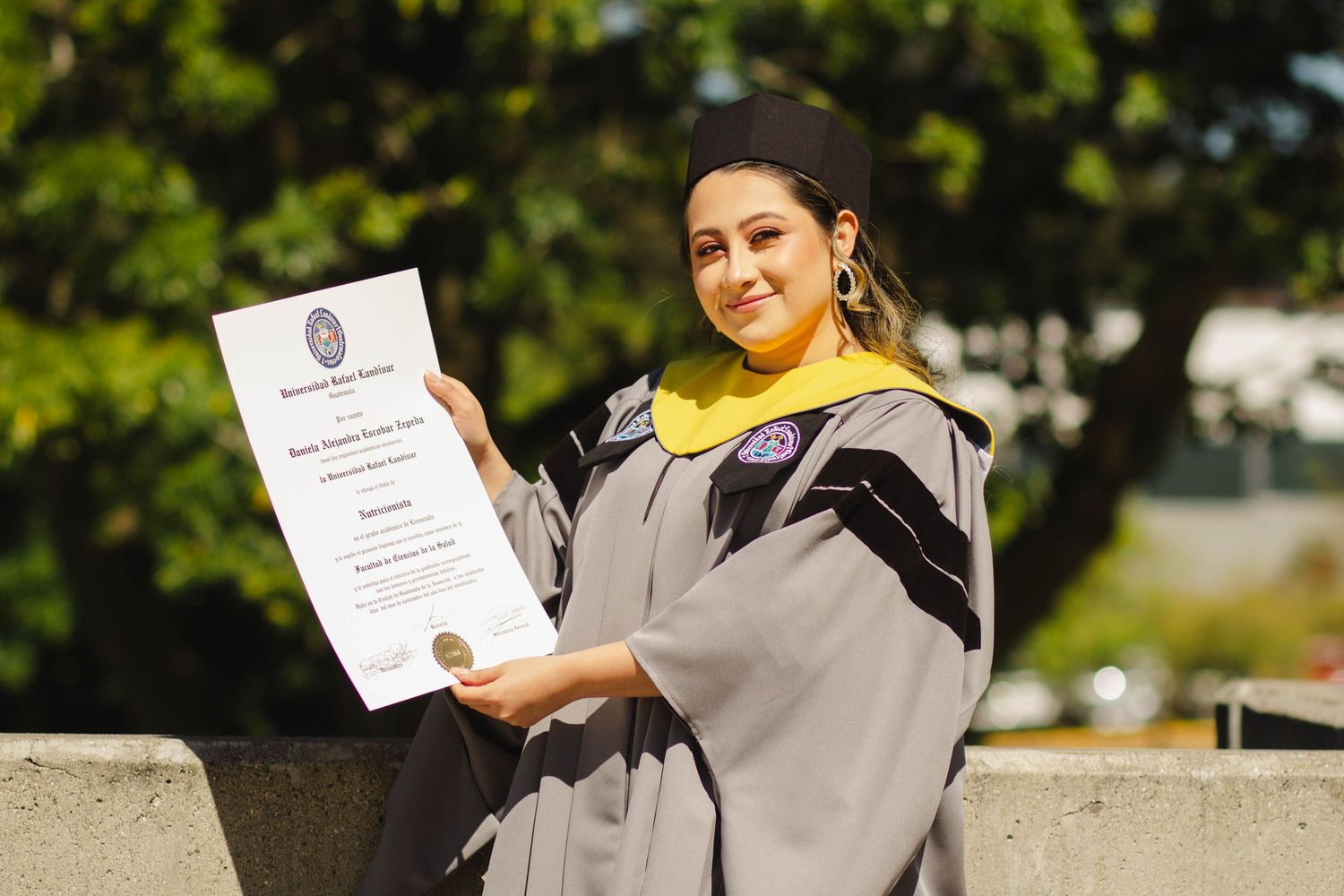 Graduate in grey academic regalia holds up diploma certificate while posing outside.