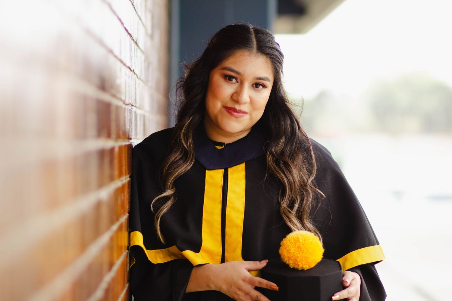 Graduate in black and gold academic regalia poses against brick wall in natural lighting.