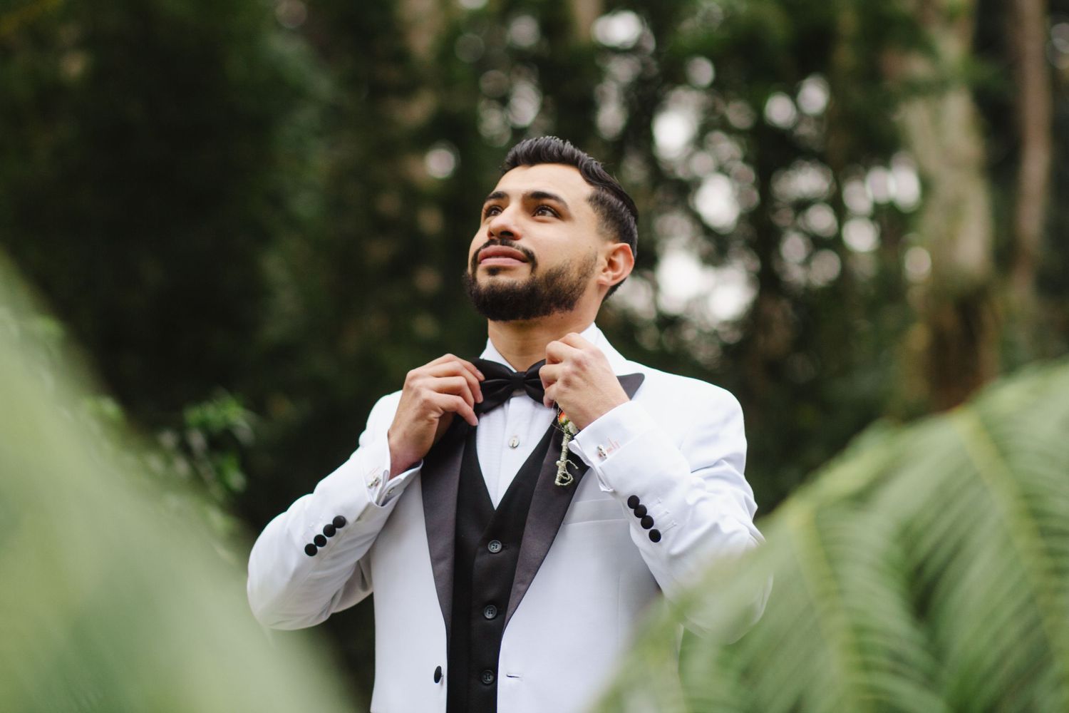 Groom in white tuxedo adjusts bowtie while surrounded by tropical greenery.