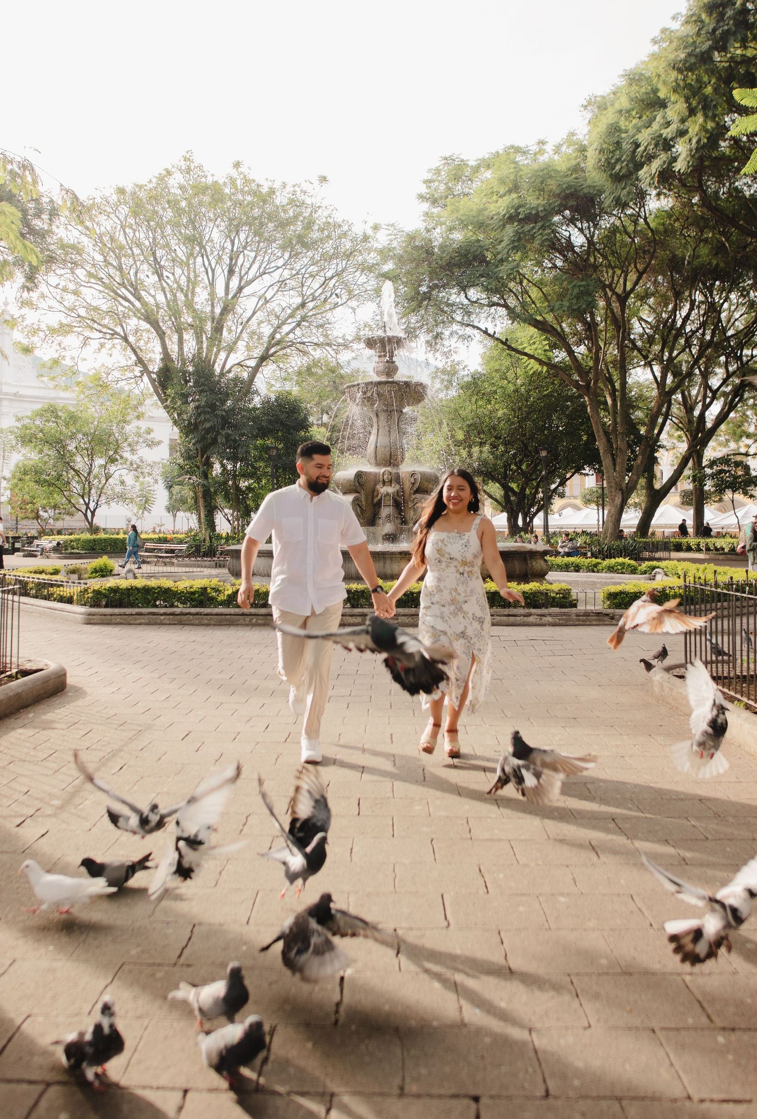 People in white outfits playfully chase pigeons in a series of photos at a park with stone pavement and trees.