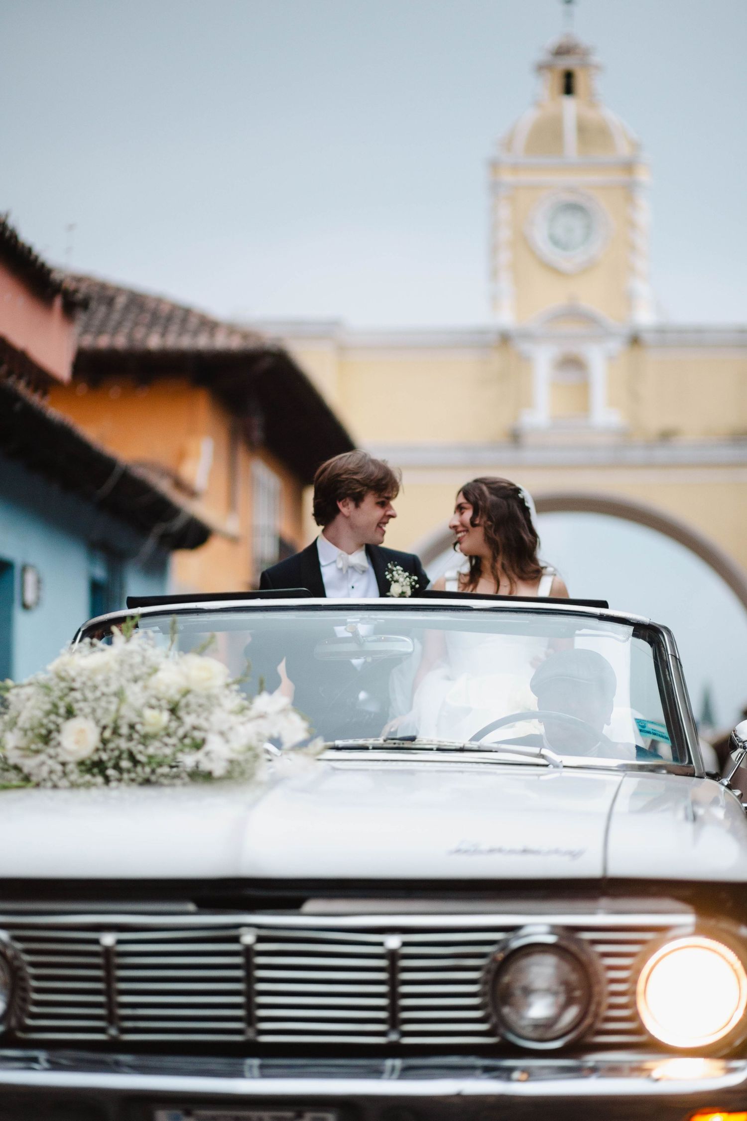 A couple in a classic convertible car during their wedding day with a colonial clock tower in the background of Antigua.