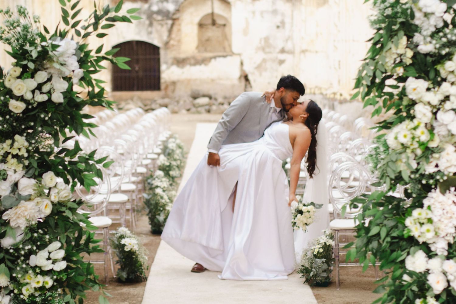 A romantic wedding couple dips in an elegant pose surrounded by white floral arrangements and stone architecture.