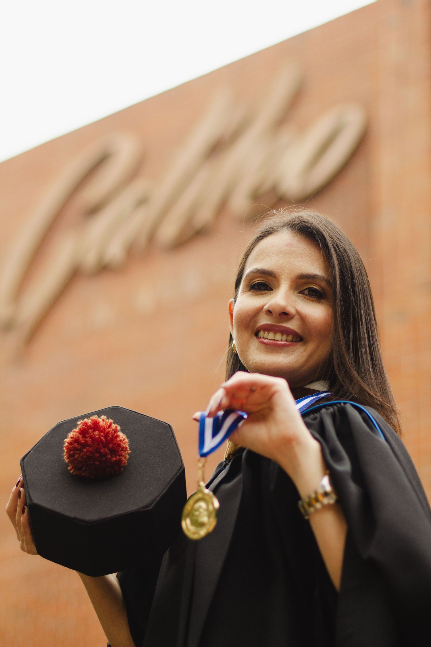 Graduate in black academic regalia holding cap with red tassel while wearing medal at graduation ceremony.