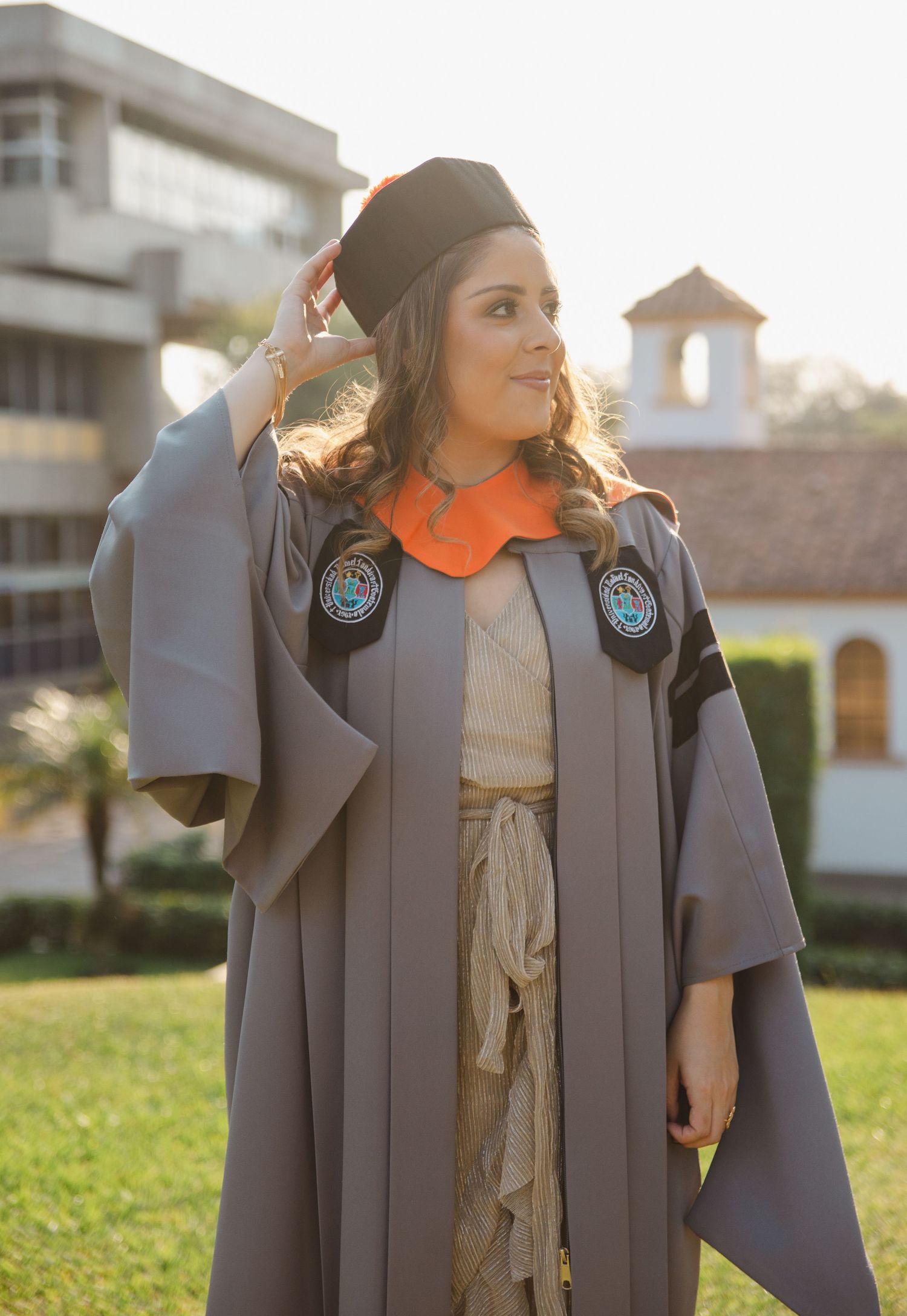 A woman in gray graduation gown and black cap smiles outdoors on a sunny campus lawn.