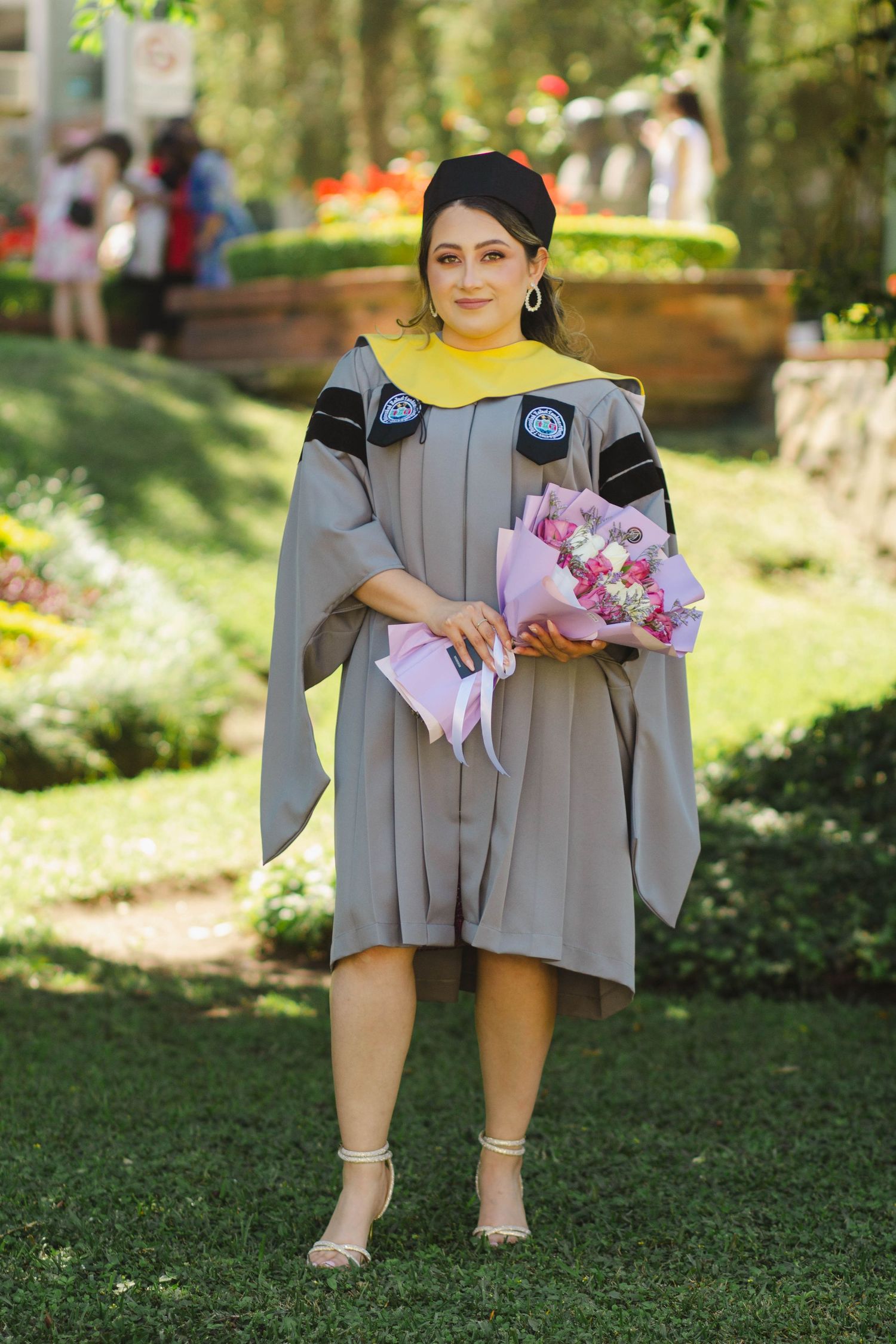 Graduate in academic regalia holding pink flowers poses on landscaped campus grounds.