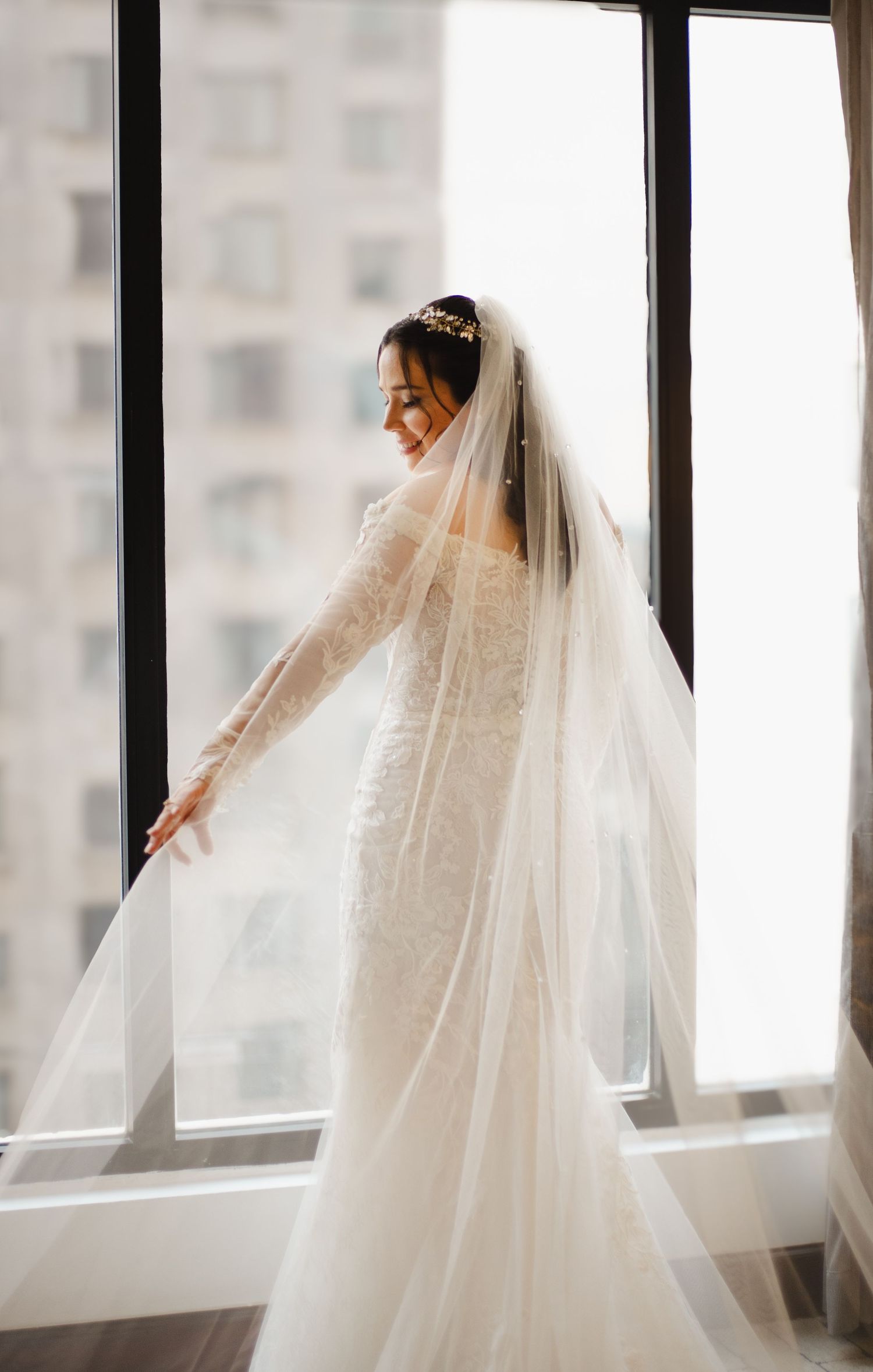 A bride in a flowing white wedding gown and long veil poses gracefully by large windows in natural light.