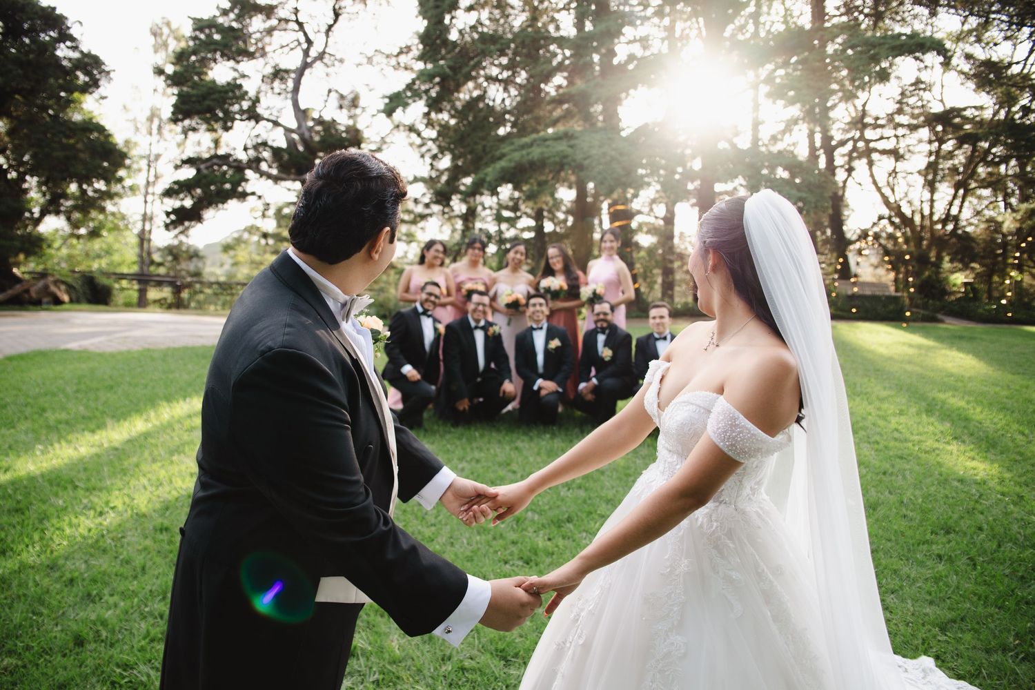 Bride and groom hold hands outdoors at sunset wedding ceremony with bridal party gathered on green lawn.