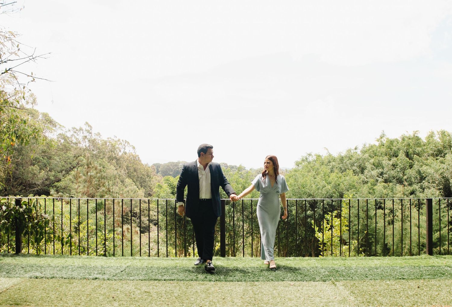 A couple walks hand in hand along a scenic overlook with lush greenery in the background on a sunny day.