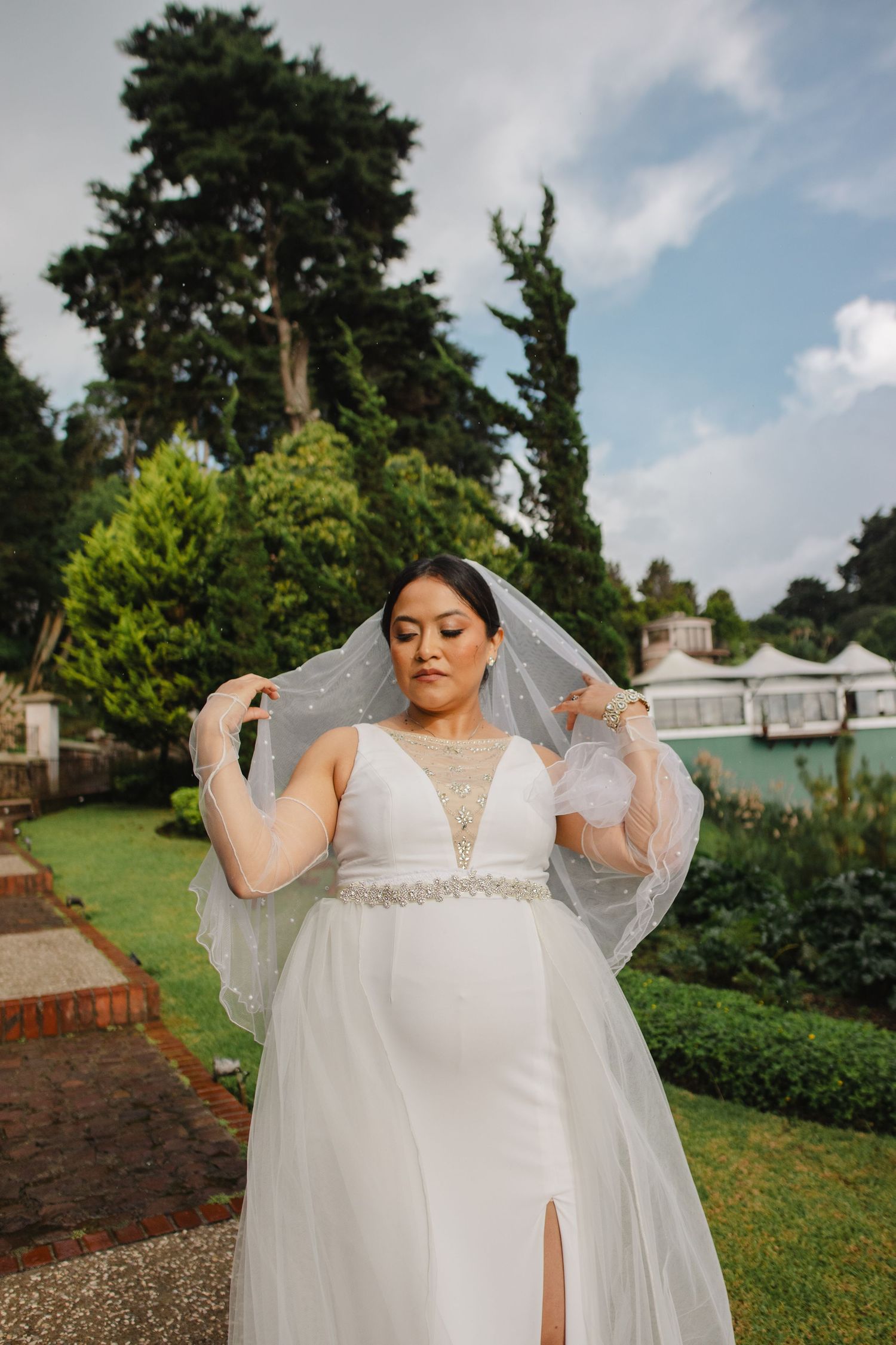A bride in a flowing white wedding dress with lace details stands in a garden setting under cloudy skies.