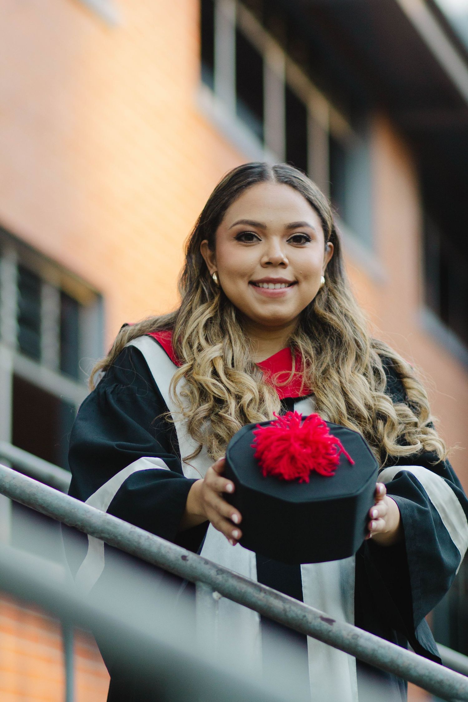 Graduate in academic dress holds graduation cap with red tassel while standing at railing.